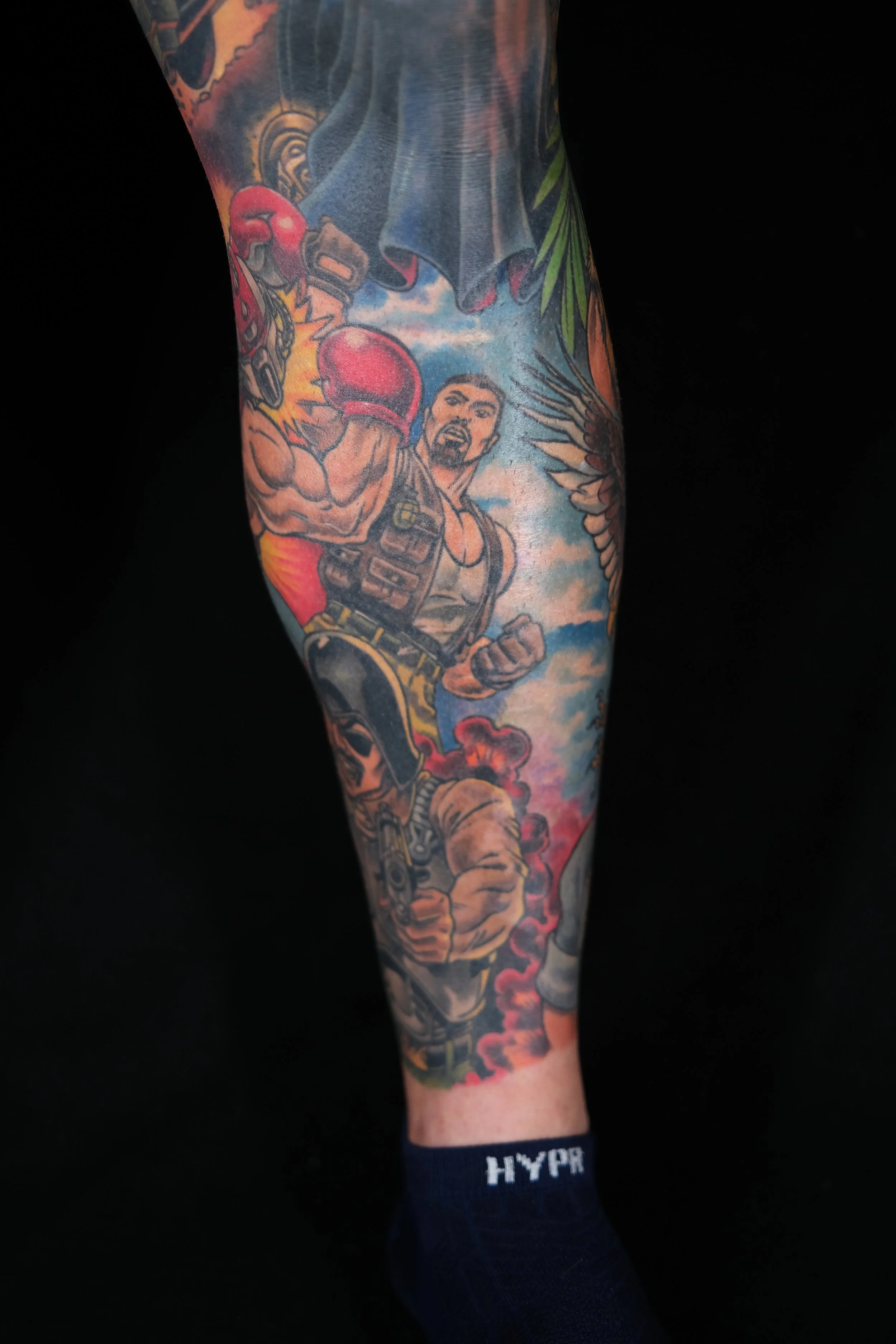 A person's arm with colorful tattoos, featuring a boxer with gloves, a woman with feathers, and a cloudy sky, wearing a black sock with the word 'HYPER' in white letters.