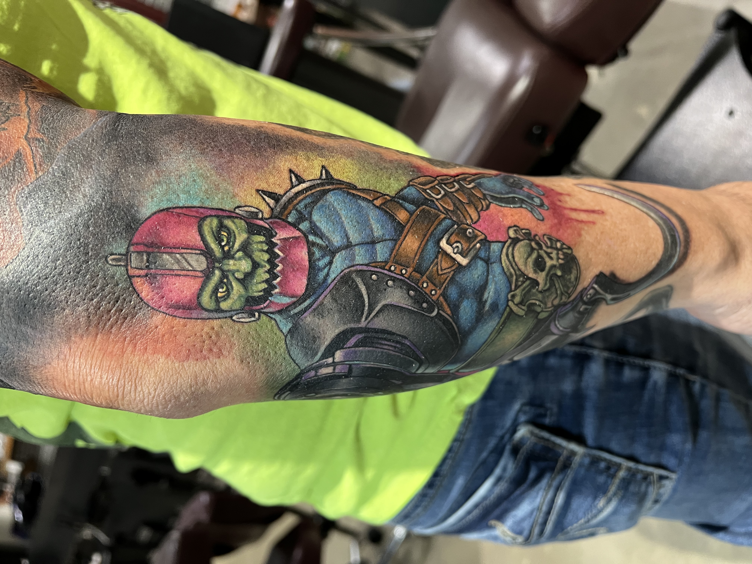 Colorful tattoo of a green skull with a pink hood, wearing a blue jacket with brown straps, on a person's arm.
