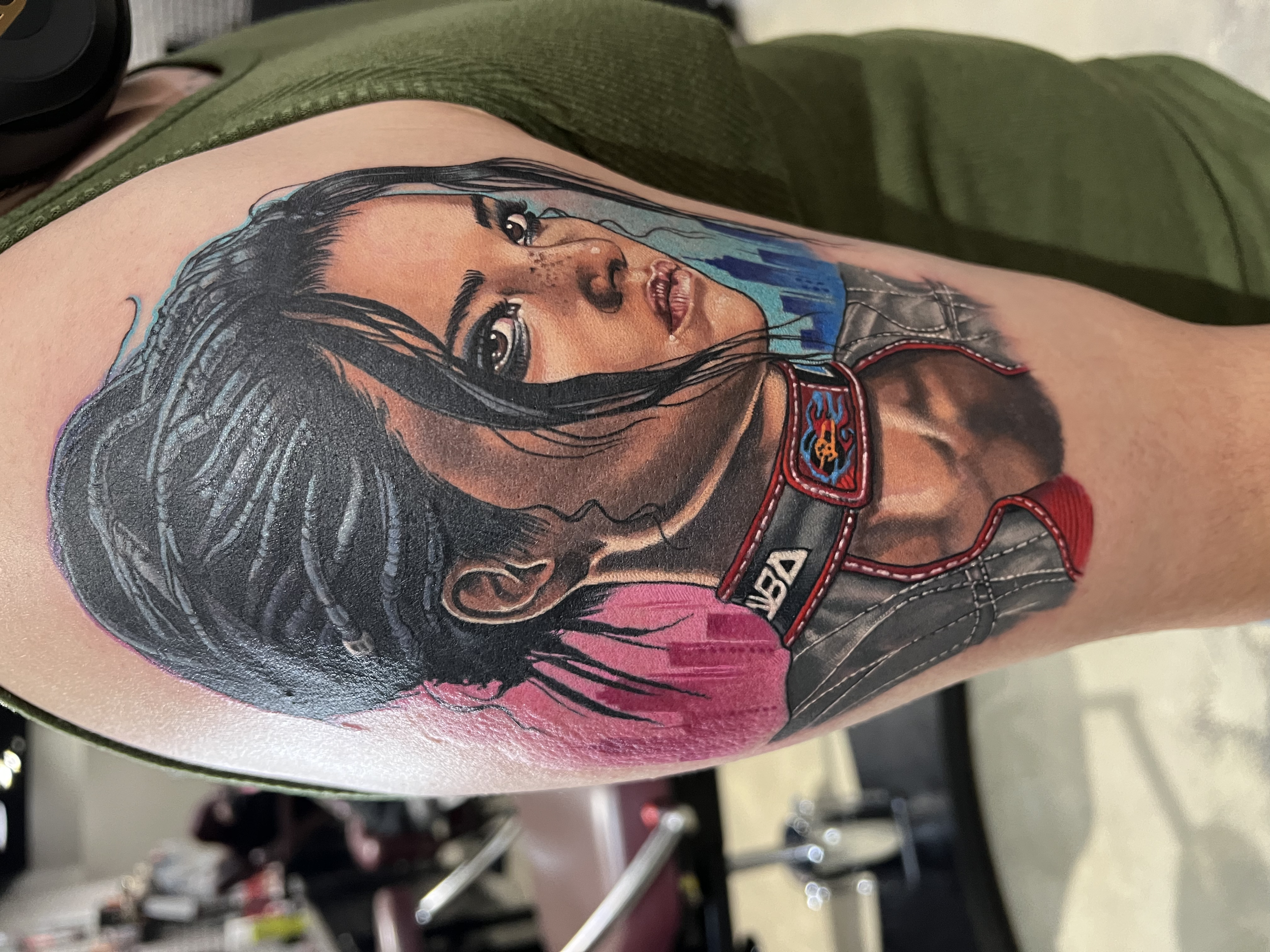Colorful tattoo of a woman's face with dark hair, on a person's upper arm. The woman has a serious expression, with detailed facial features and a neck tattoo. She is wearing a blue scarf or shirt.