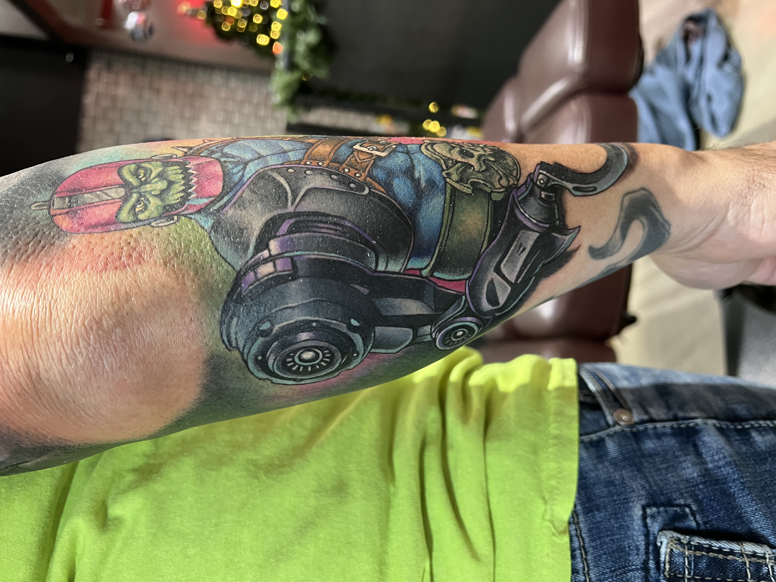 Close-up of a person's colorful tattooed arm and hand, with a background including a brown leather chair, blue jeans, and a blurred Christmas decoration with lights.