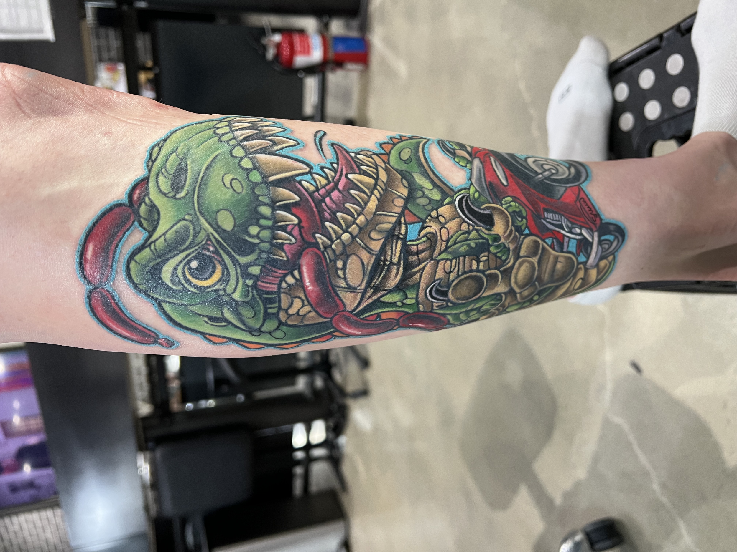 Colorful tattoo of a t-rex riding a motorcycle with sausages in his mouth.