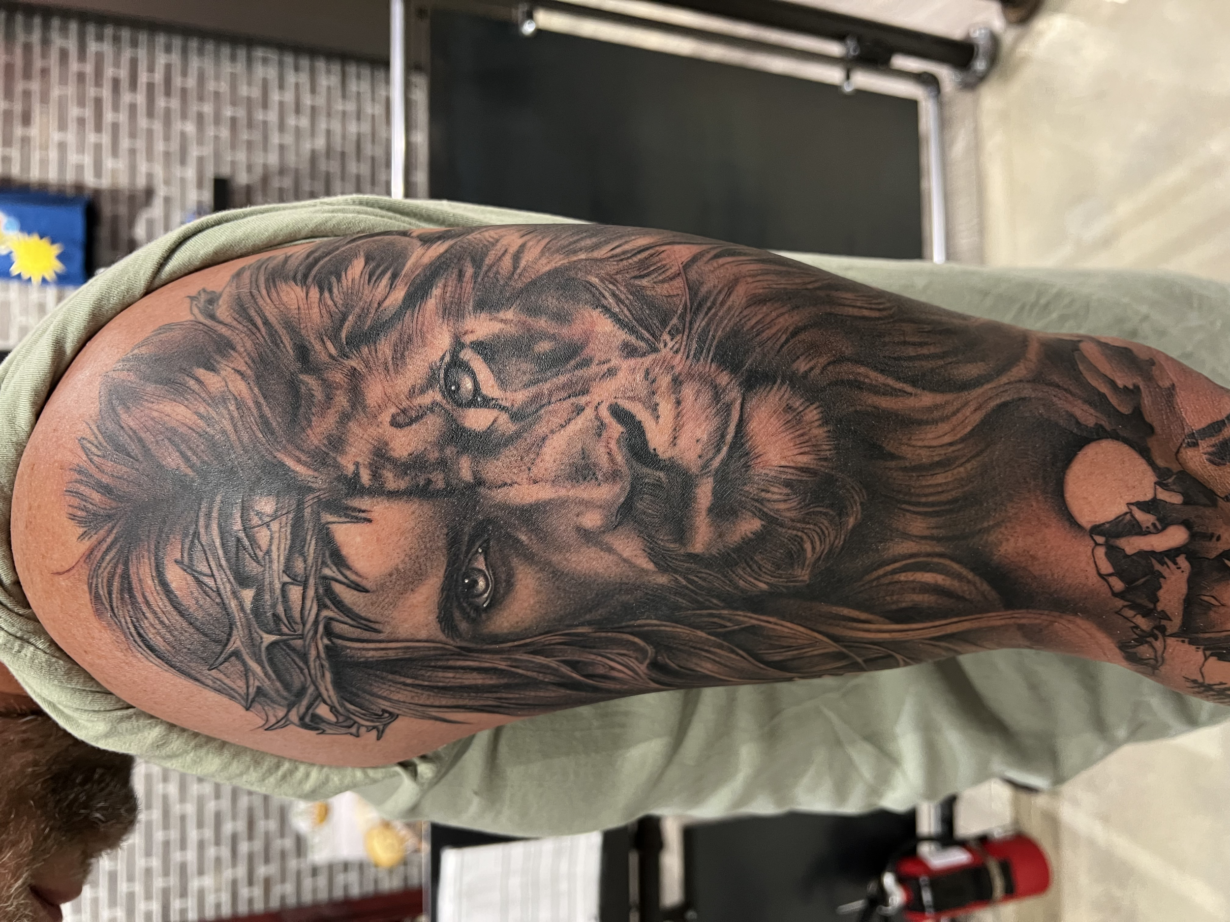 Close-up of a person's upper arm with a detailed black and gray tattoo of a lion's face surrounded by mane.