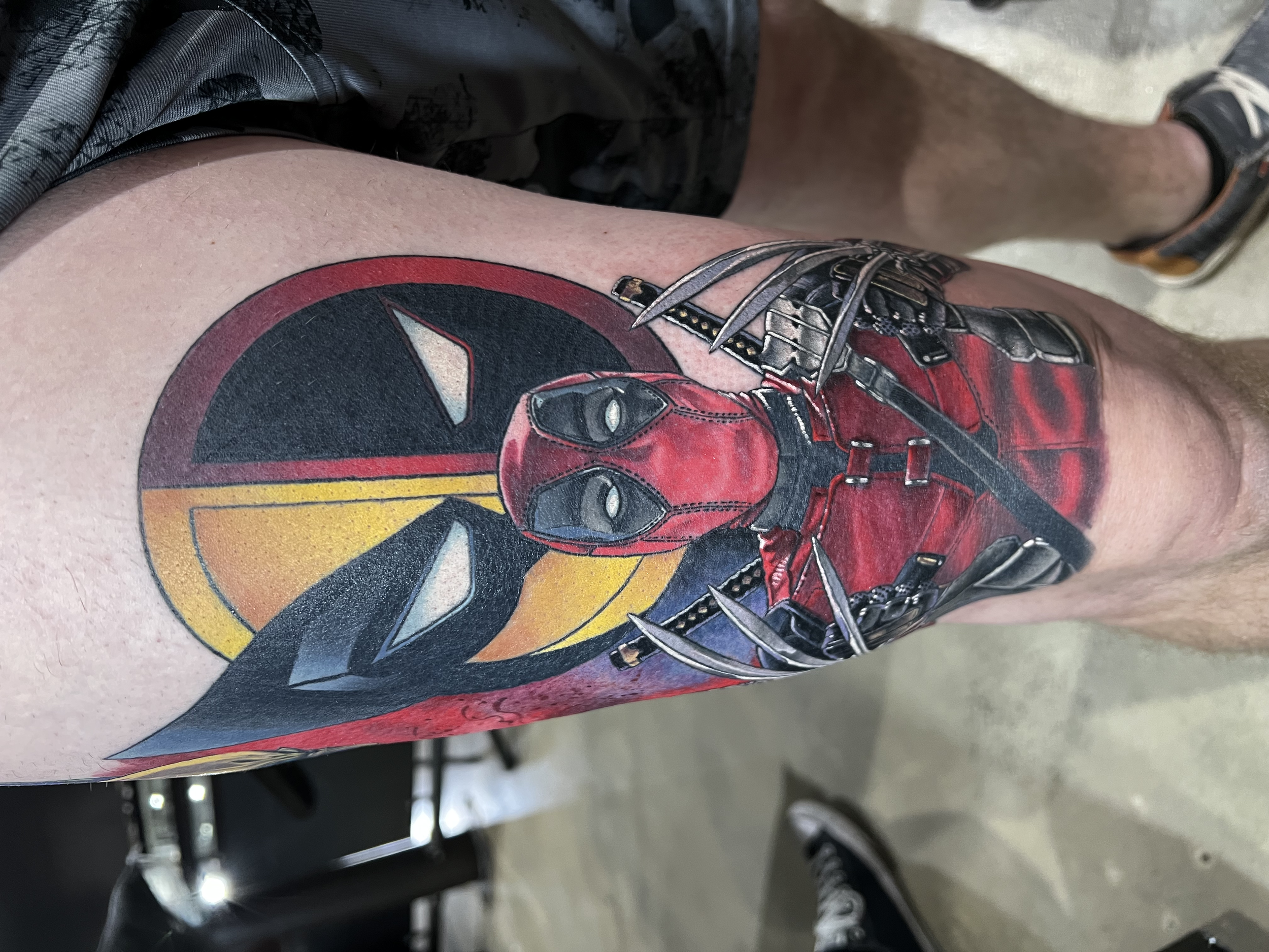 Close-up of a person's thigh with Deadpool tattoo, wearing black and red sneakers, standing on a concrete floor.
