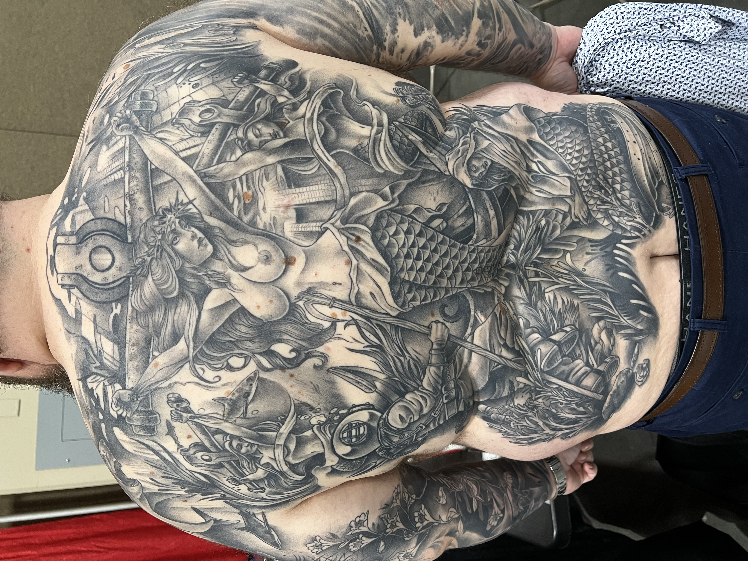 A person with extensive black and gray tattoos covering their upper body, including a detailed depiction of a woman with flowing hair, armor, and a dragon. The person is wearing a white shirt with blue patterns and dark blue pants with a brown belt.