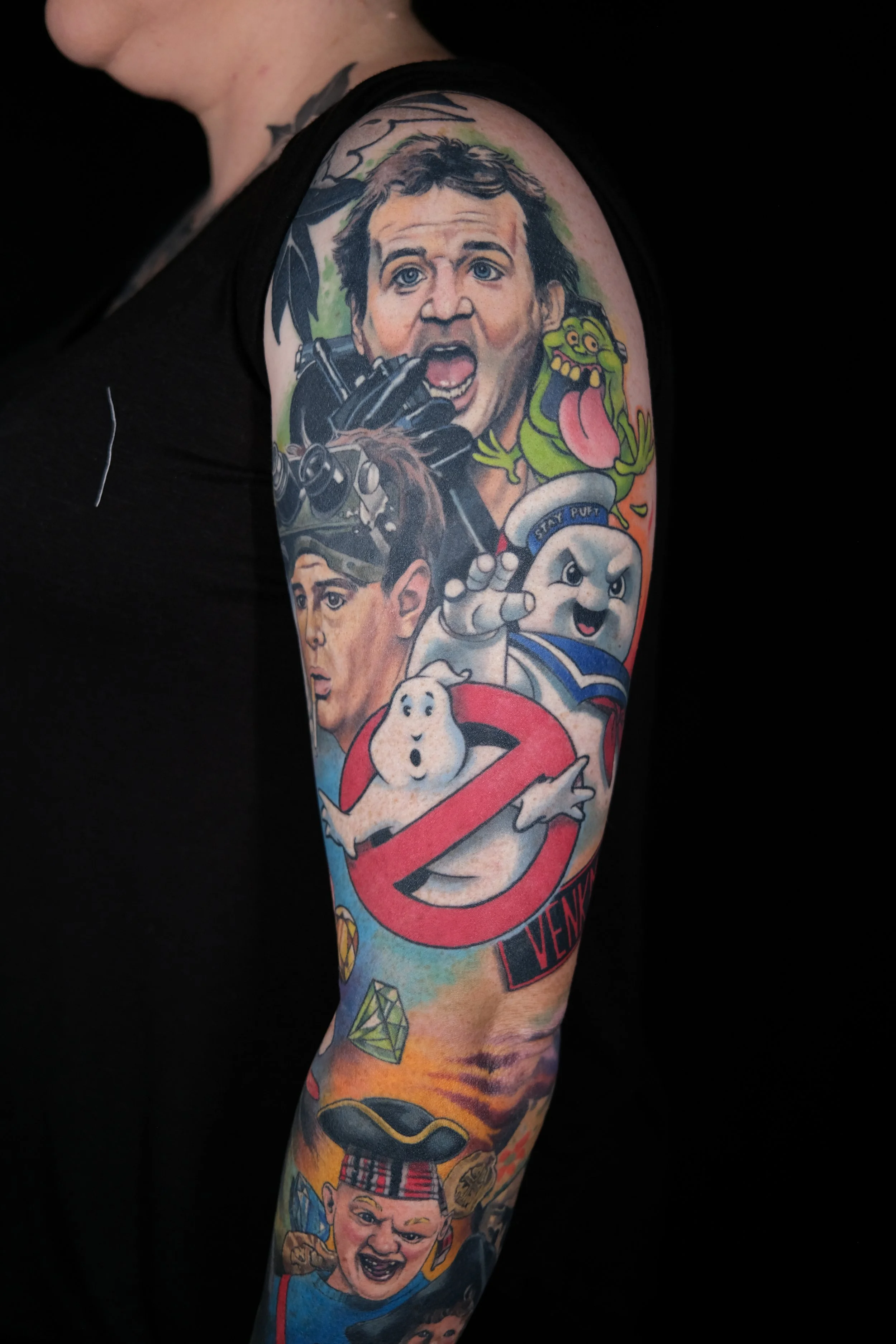 Colorful tattoo on arm featuring characters from Ghostbusters, including a woman with blonde hair, a green ghost, a boy with a backpack, and a ghost holding a red circle with a line through it.