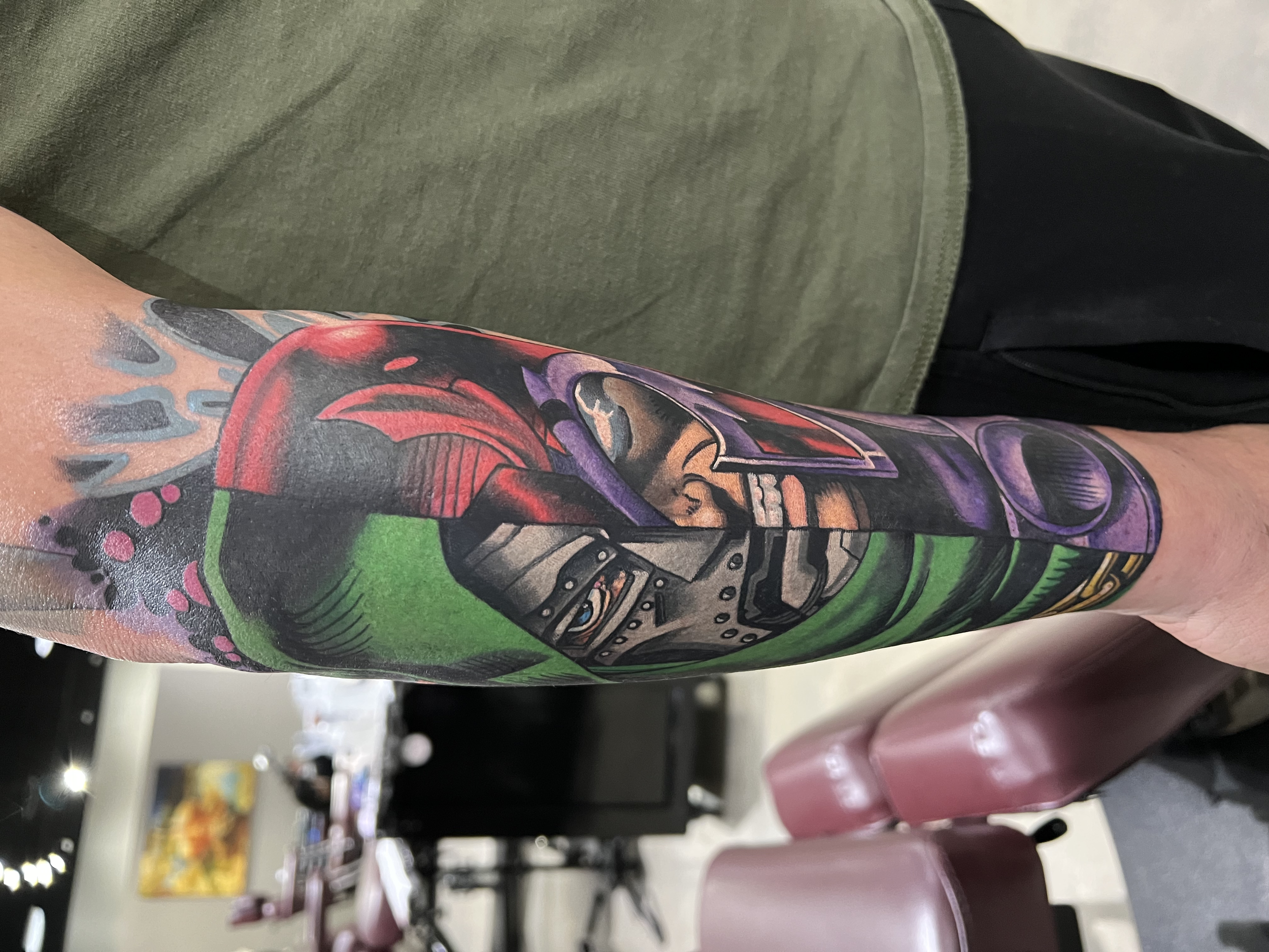Close-up of a person's arm with colorful tattoo art, showing a robotic figure and a face with closed eyes, with blurred supermarket shopping carts in the background.