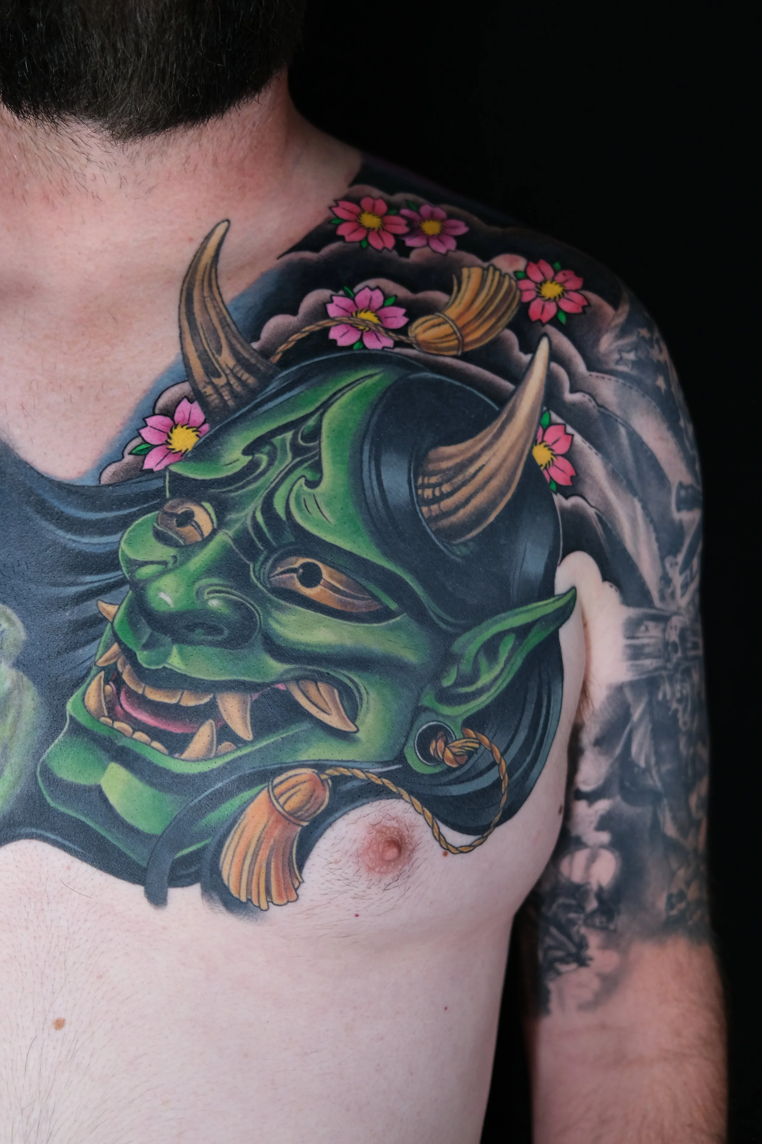 Colorful tattoo of a green Oni mask with horns, pink cherry blossoms, and clouds on a man's shoulder and chest.
