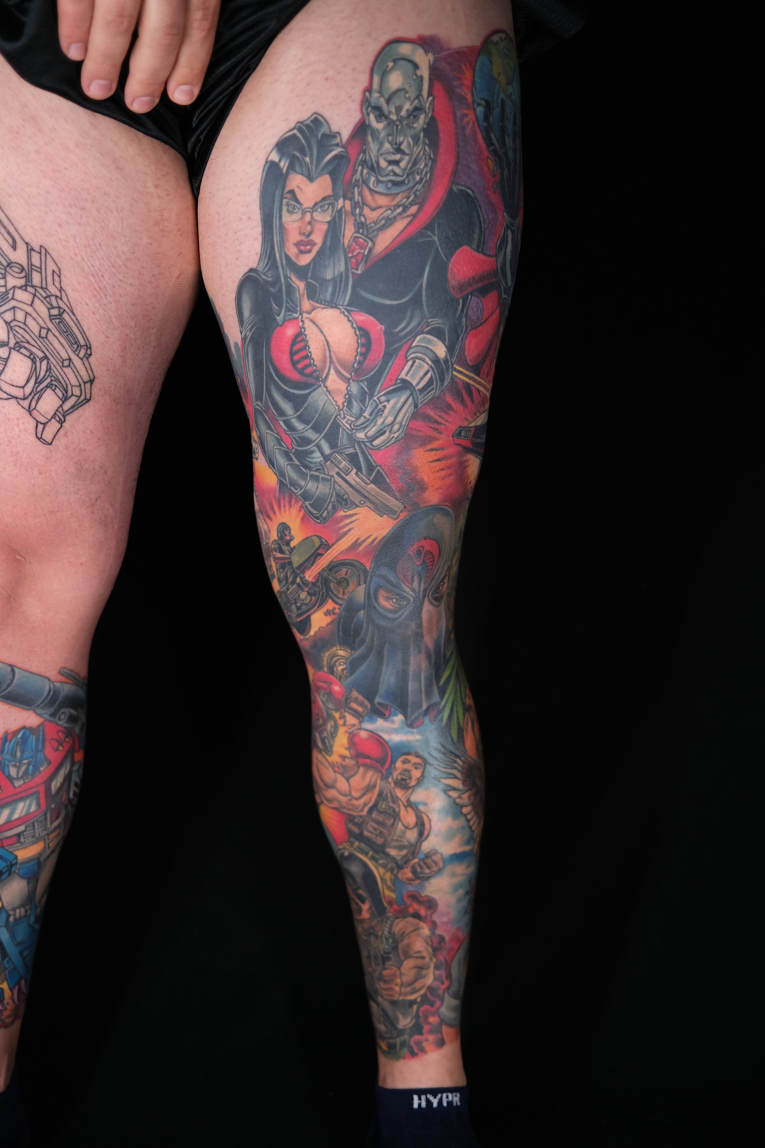 Colorful tattoo on a person's thigh depicting a woman with glasses and a revealing outfit, and a large muscular man with a chain around his neck, surrounded by explosions, vehicles, and action scenes.