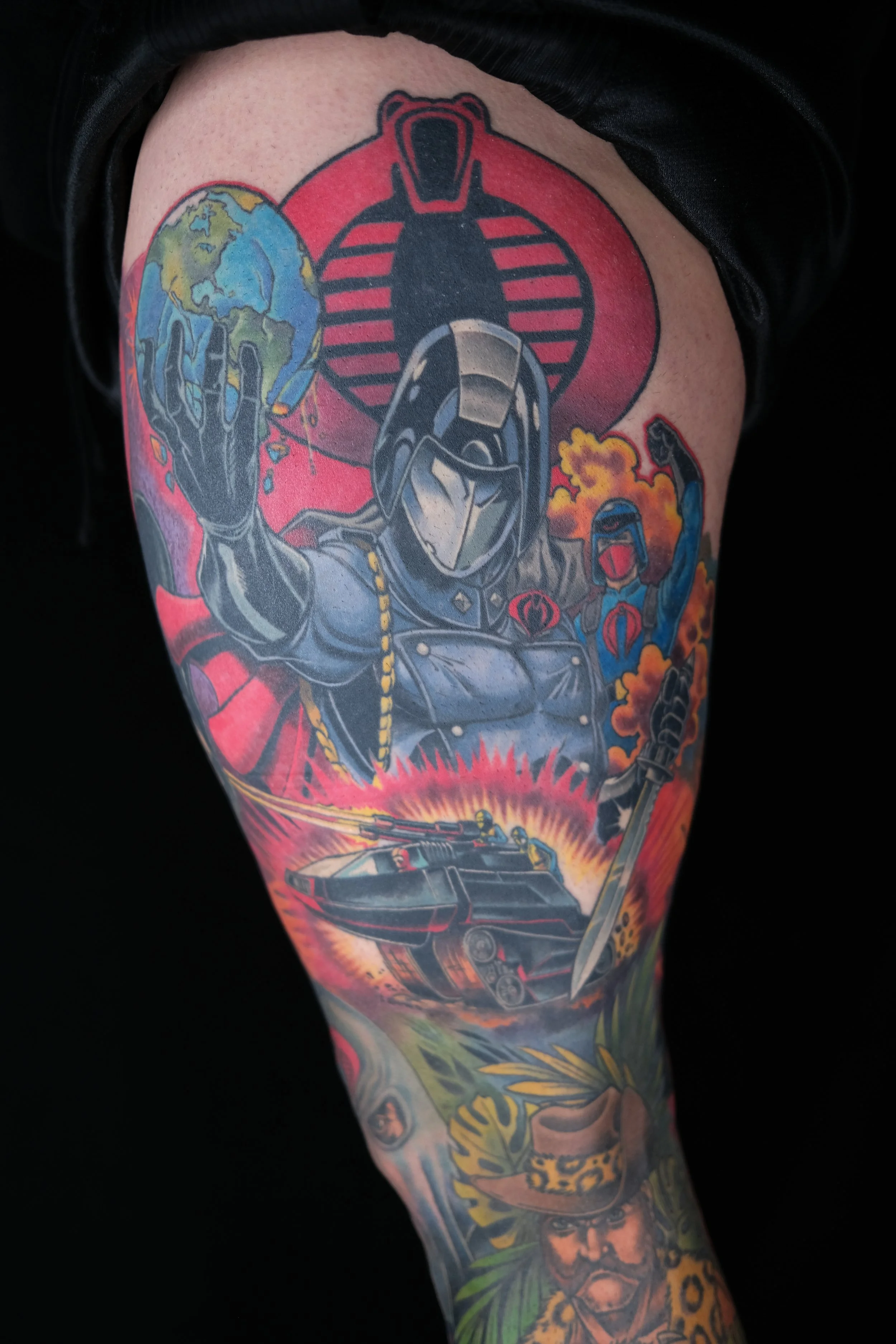 Tattoo of a soldier in armor with a tank, a masked figure, and a burning background, with a globe on the hand, and a Cobra emblem in the background.