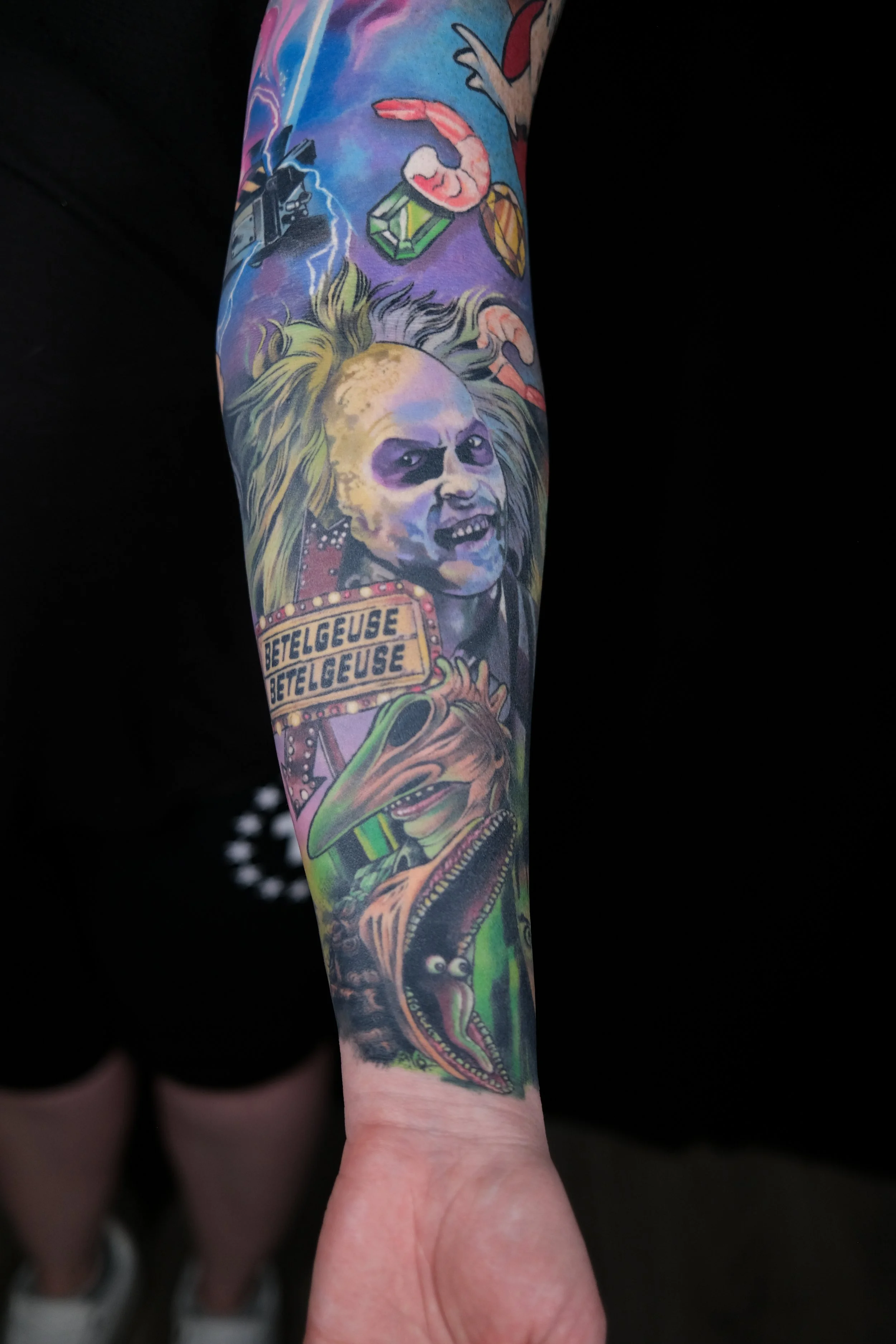 Colorful tattoo sleeve featuring a skeleton with wild hair, a crocodile, a woman with an expression, and text reading 'BETELGEUSE' repeatedly.