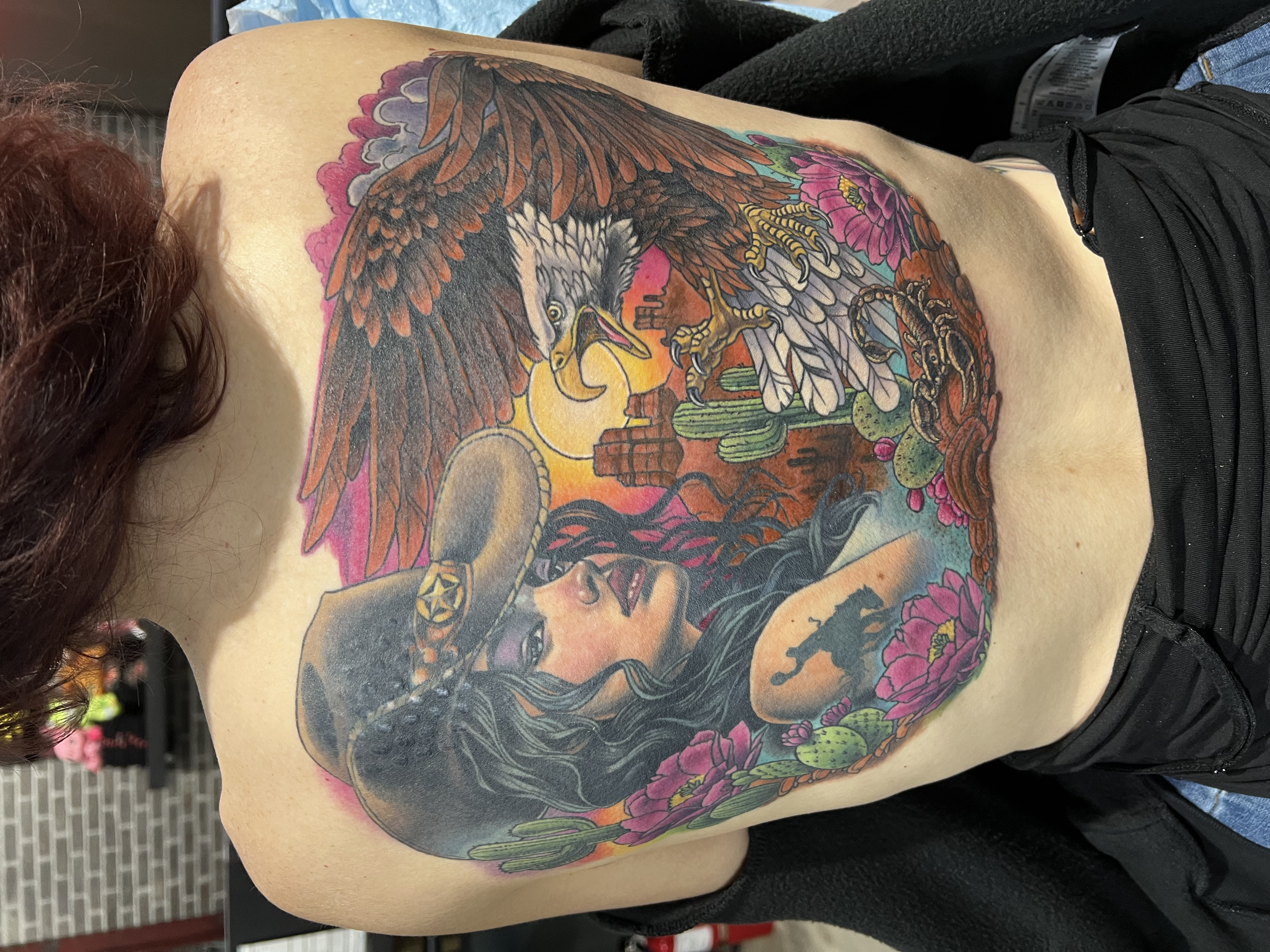 A detailed colorful tattoo of an eagle with outstretched wings, perched on a cactus, surrounded by vibrant pink flowers, with a sunset or sunrise background featuring an orange and yellow sun. Tattoo Covering up non-laser tattoo removal scars.