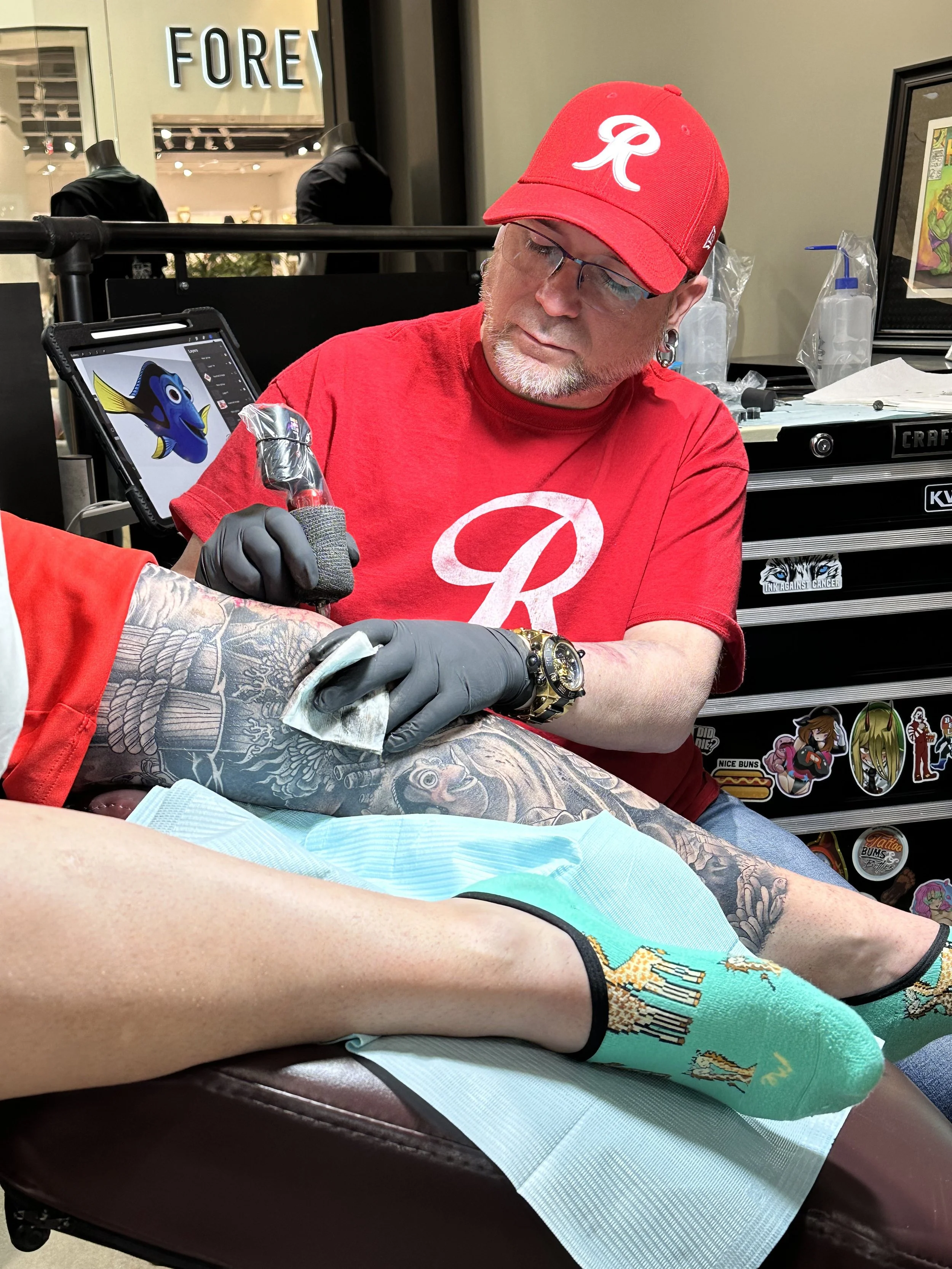 A person with tattoos on their leg lying on a padded surface while a tattoo artist in a red shirt and cap with a white logo tattoos their leg. The tattoo artist is wearing black gloves and glasses, and is using a tattoo machine. The background shows a toolbox with stickers, a bottle of disinfectant, and a framed artwork.