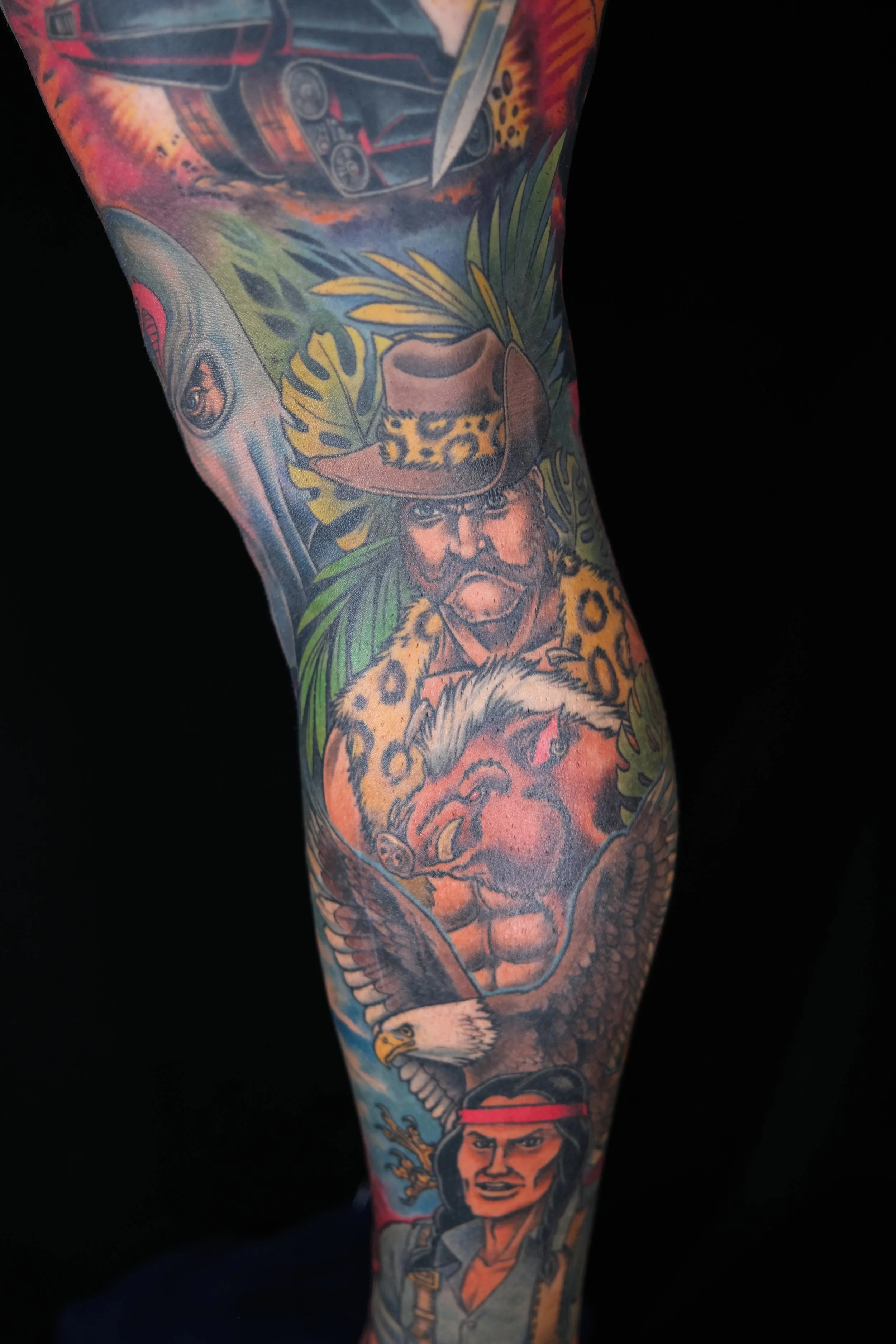 A colorful tattoo sleeve featuring various pop culture characters and elements, including a portrait of a woman with dark hair and a confident expression, surrounded by animals and objects like a leopard, an eagle, and a cowboy hat.