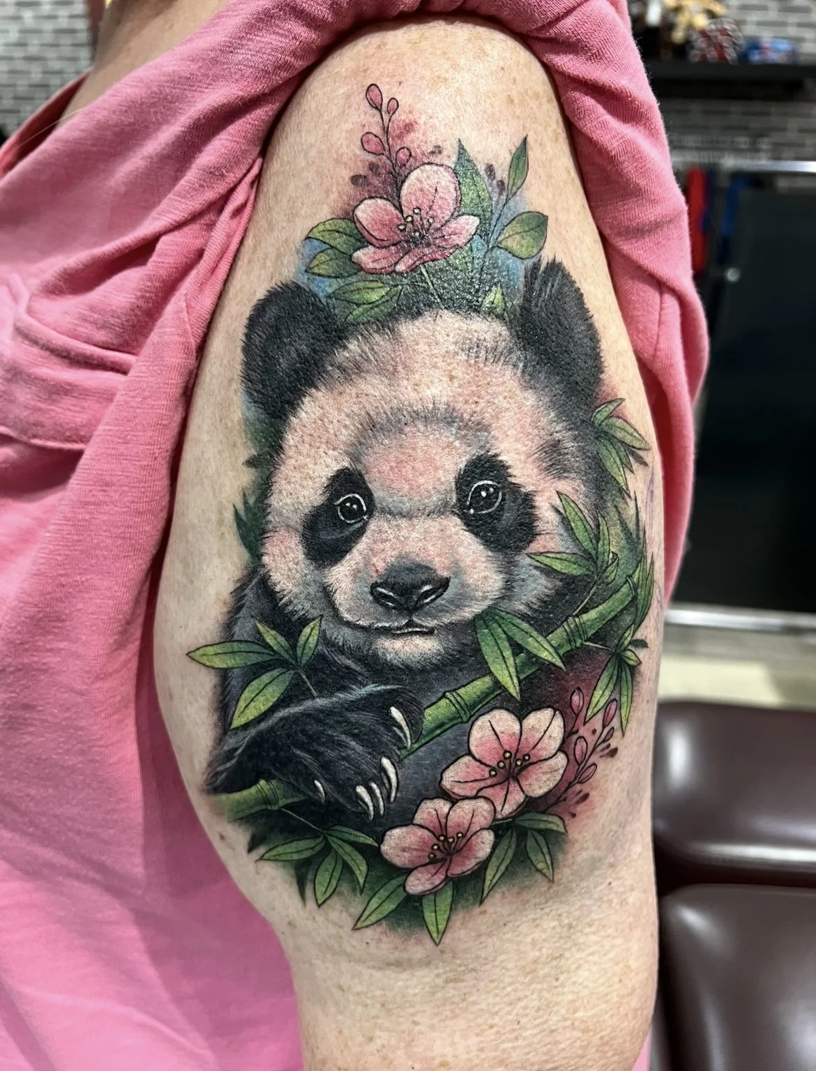 Colorful tattoo of a baby panda holding a bamboo stalk, surrounded by pink flowers and green leaves on a person's upper arm.