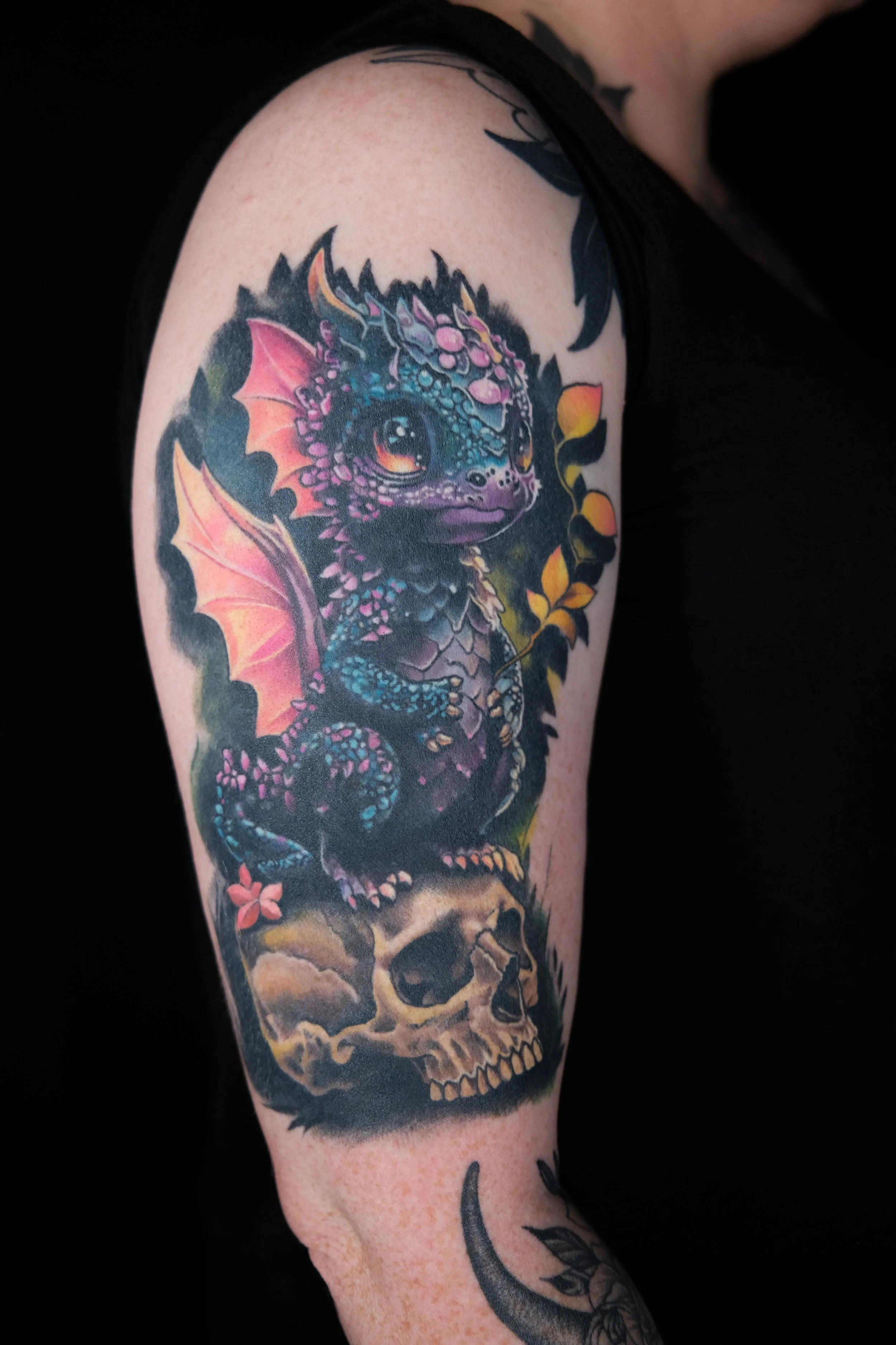 Colorful tattoo of a dragon with wings and a skull, accented with flowers and foliage on a person's upper arm.