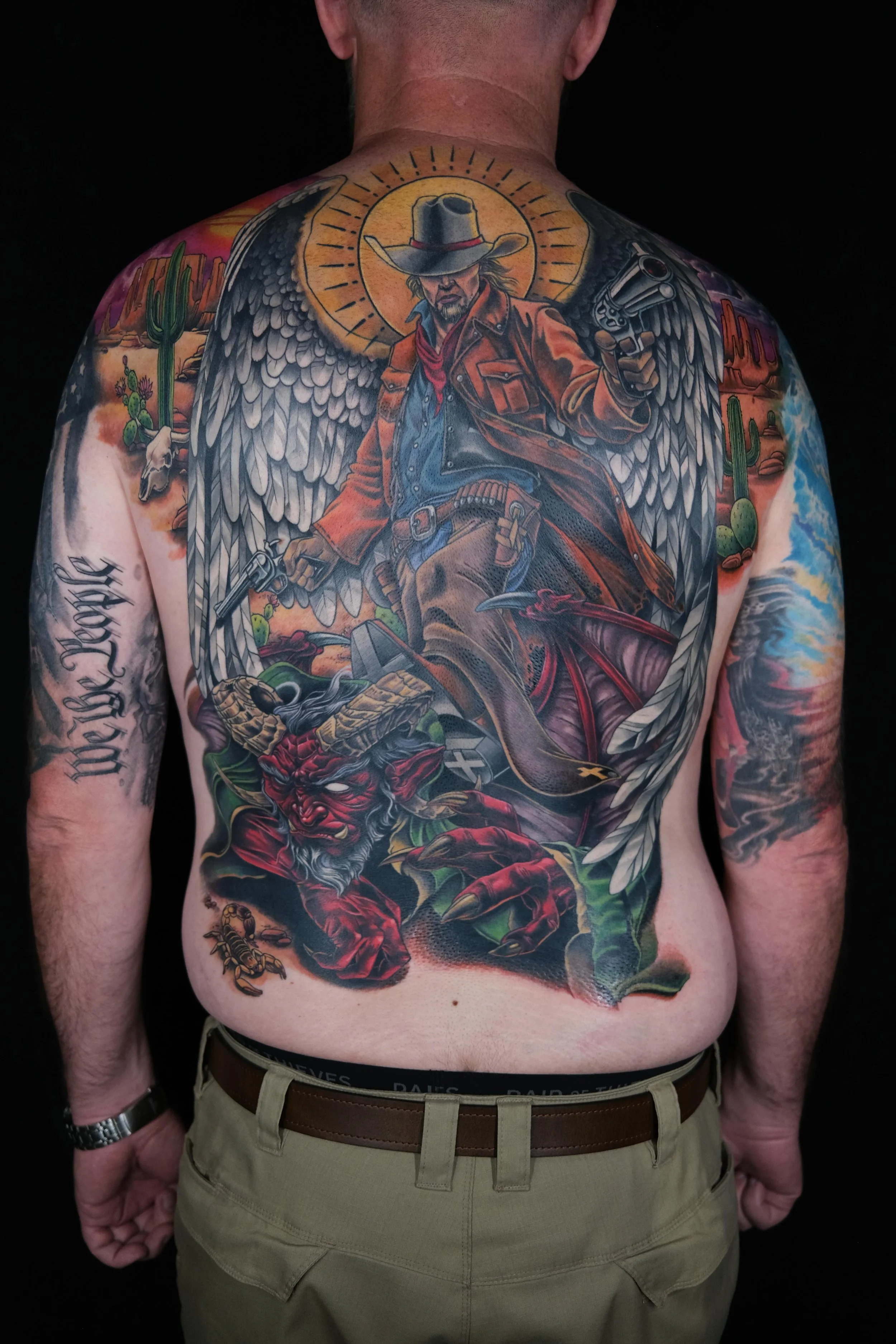 Colorful tattoo on a man's back depicting a cowboy with wings, holding a revolver, with desert landscape, cacti, mountains, and a red devil figure below.