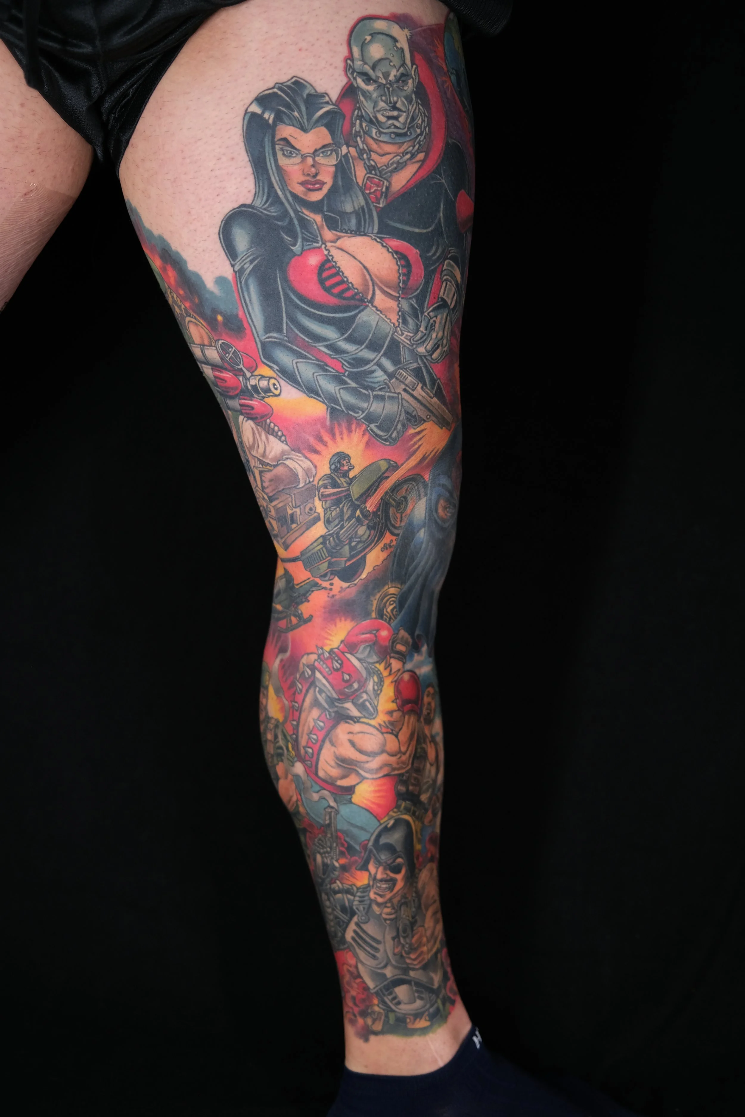 Full-length sleeve tattoo on a person's arm featuring a woman with glasses and long dark hair wearing leather clothing, a muscular man with a mohawk and chains, and other various characters and scenes in a colorful and detailed style.