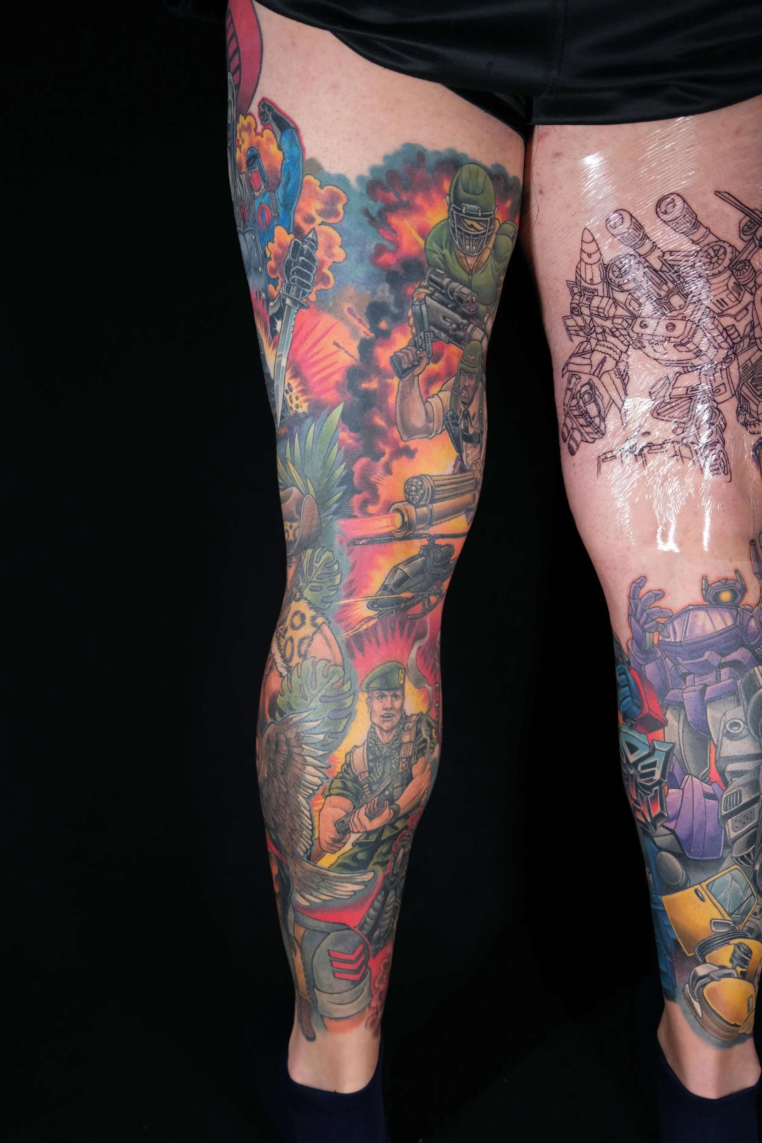 Colorful tattoo sleeve featuring soldiers, explosions, and military themes on person’s legs, with a black background.