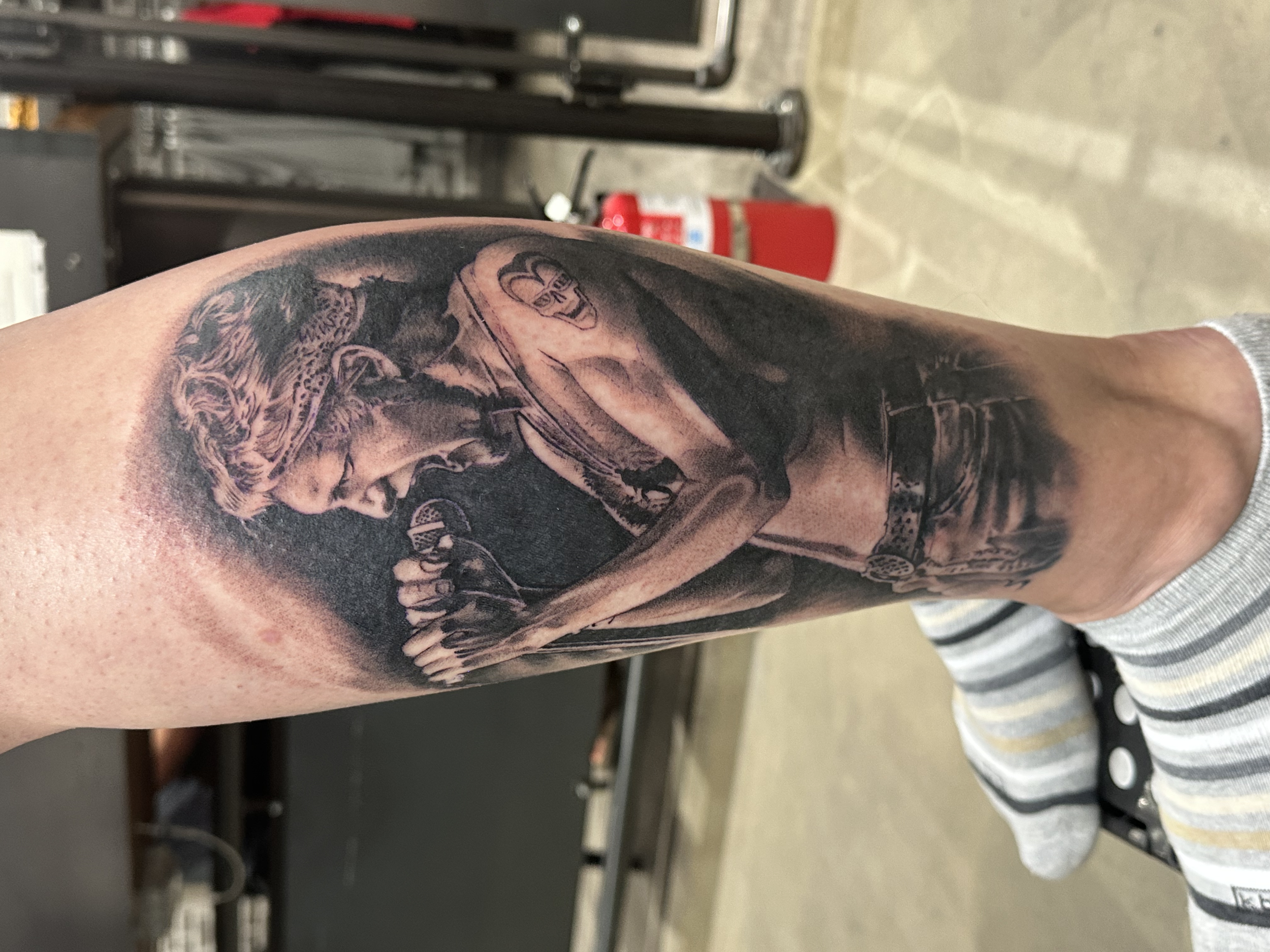 A tattoo of a person's face, wearing sunglasses and a cap, with detailed shading and realistic style. The tattoo is located on the person's upper arm.