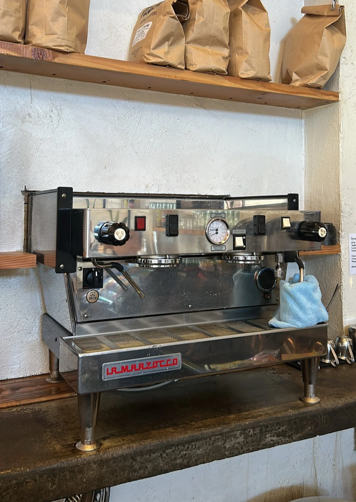 A stainless steel espresso machine with black and red controls placed on a wooden counter, with paper bags on a shelf above it and a cloth hanging on the right side.