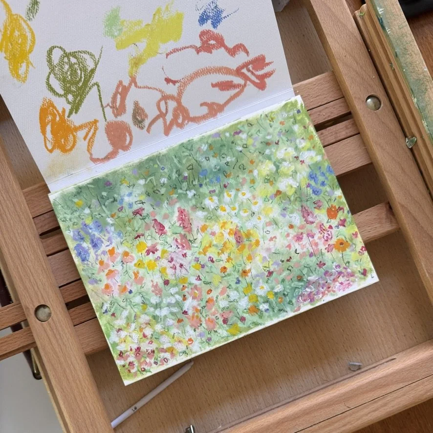 Children's colorful crayon drawing of animals on white paper and a painting of a field of wildflowers on watercolor paper, with a wooden art easel.