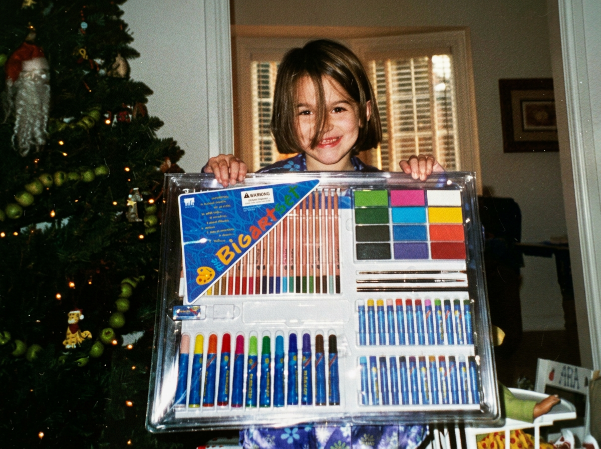 A young girl with short brown hair smiling and holding a large set of colorful art supplies in front of a decorated Christmas tree inside a house.