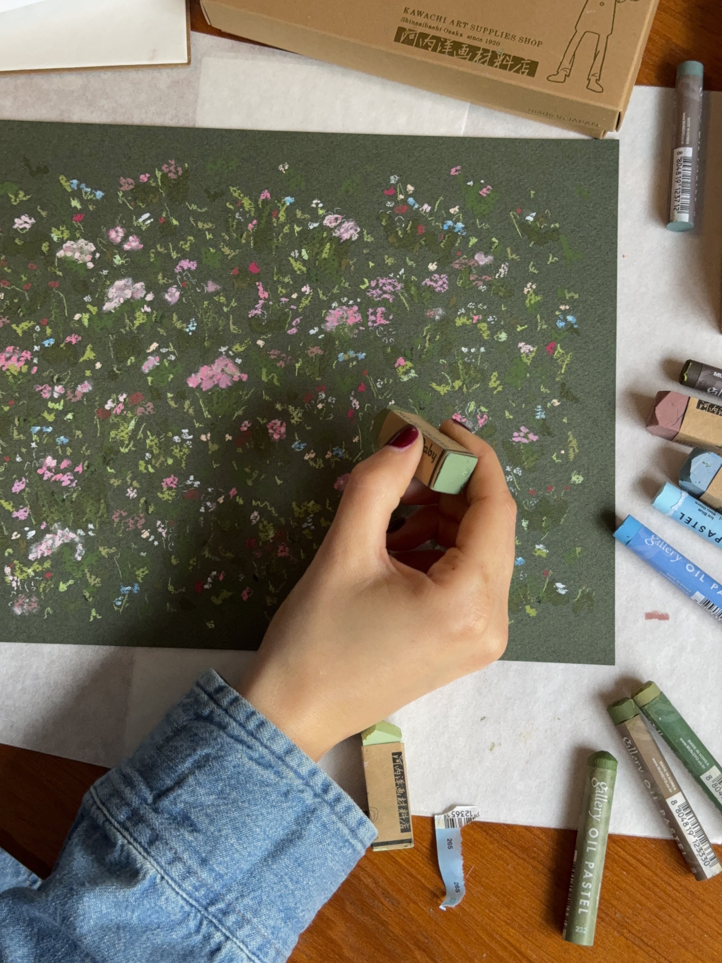 Person with painted nails holding a pastel green oil pastel, surrounded by various pastel oil pastels on a wooden table, including a green one labeled 'oil pastel' and a blue one labeled 'Palette'. A green textured paper with a colorful floral print is in the center, and a box labeled 'Kawachi Art Supplies Shop' is visible at the top.