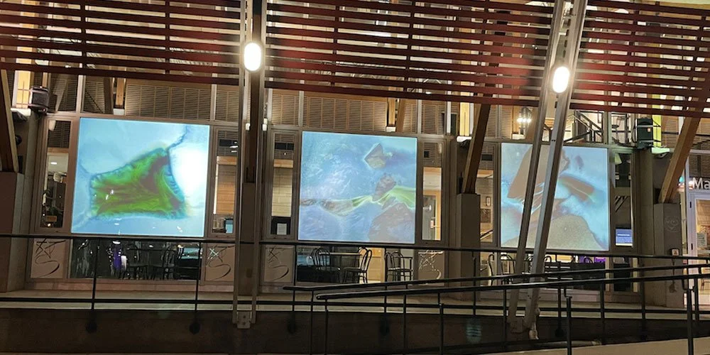 Three illuminated screens displaying abstract, colorful images in a modern indoor space with wooden and metal architectural elements.