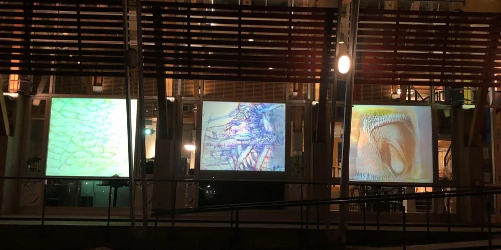 Three large screens displaying abstract images in a dark indoor setting with wooden panel ceiling and metal railings.