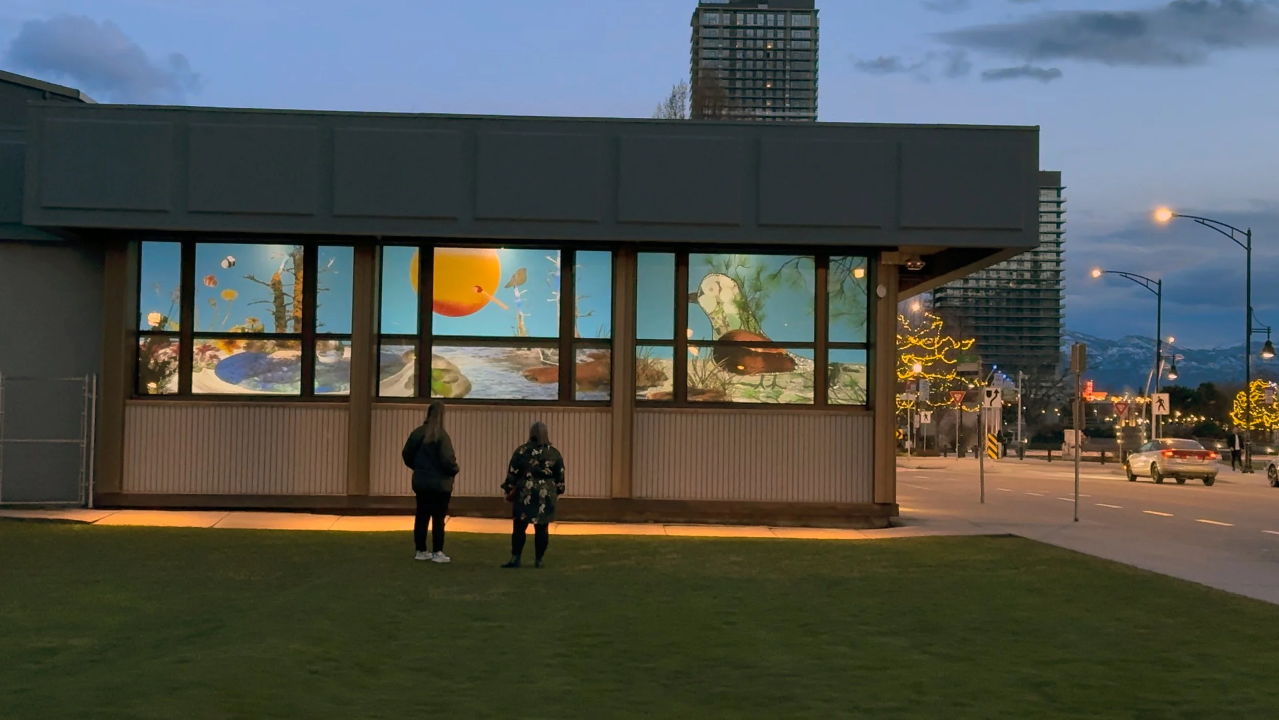 Two people stand on grass outside a building with large illuminated windows displaying a colorful mural of nature scenes, with city streets and mountains in the background at dusk.