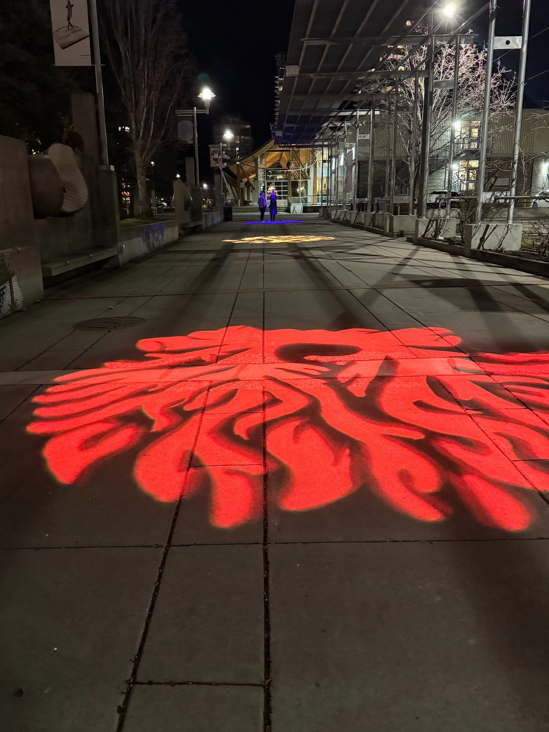 Nighttime sidewalk with illuminated red graphic of a lion's face projected on the ground, with two people walking in the distance and a building with a glass entrance.