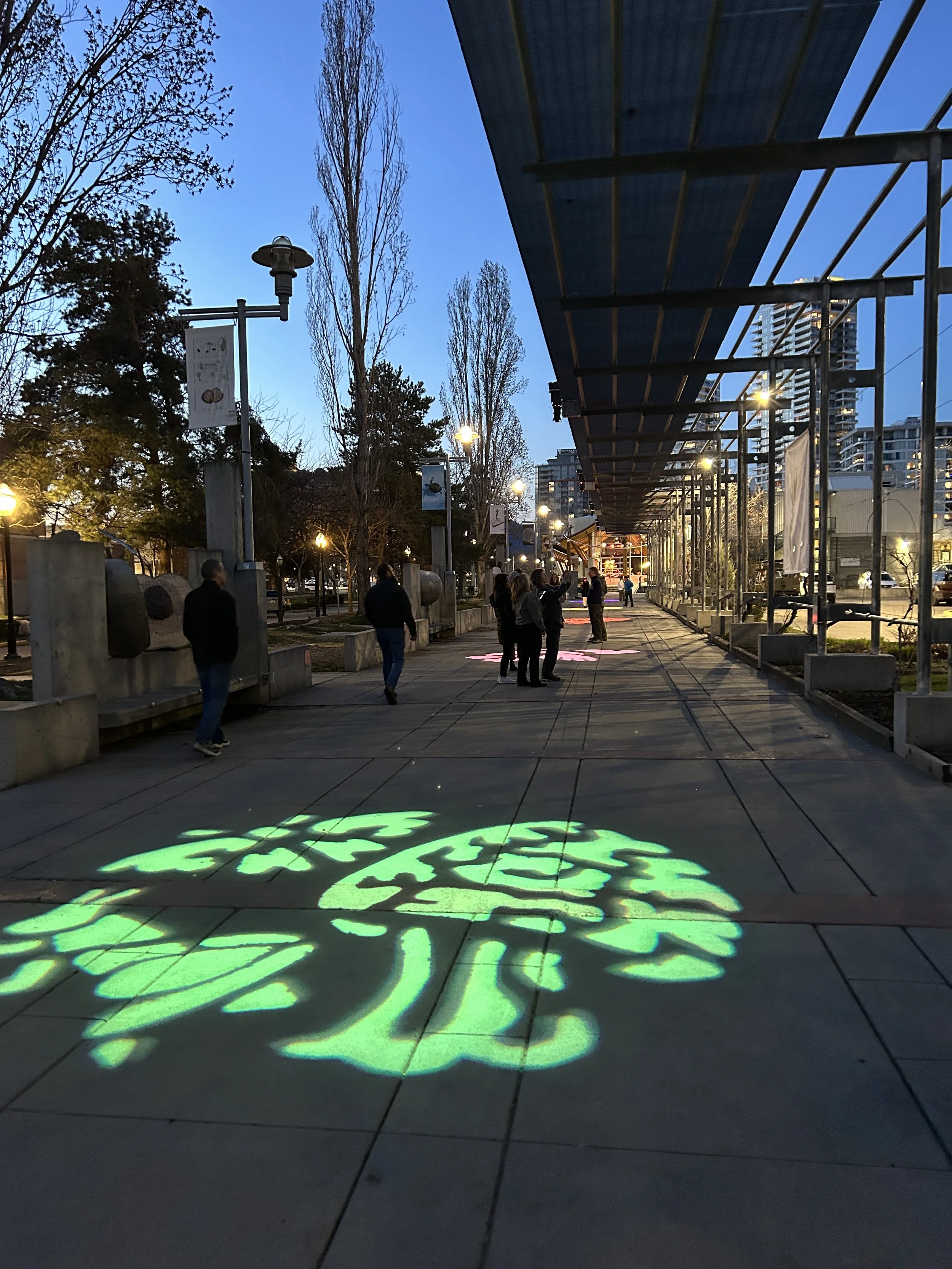 People walking along a city sidewalk with illuminated green and pink projections on the ground and streetlights glowing at dusk.