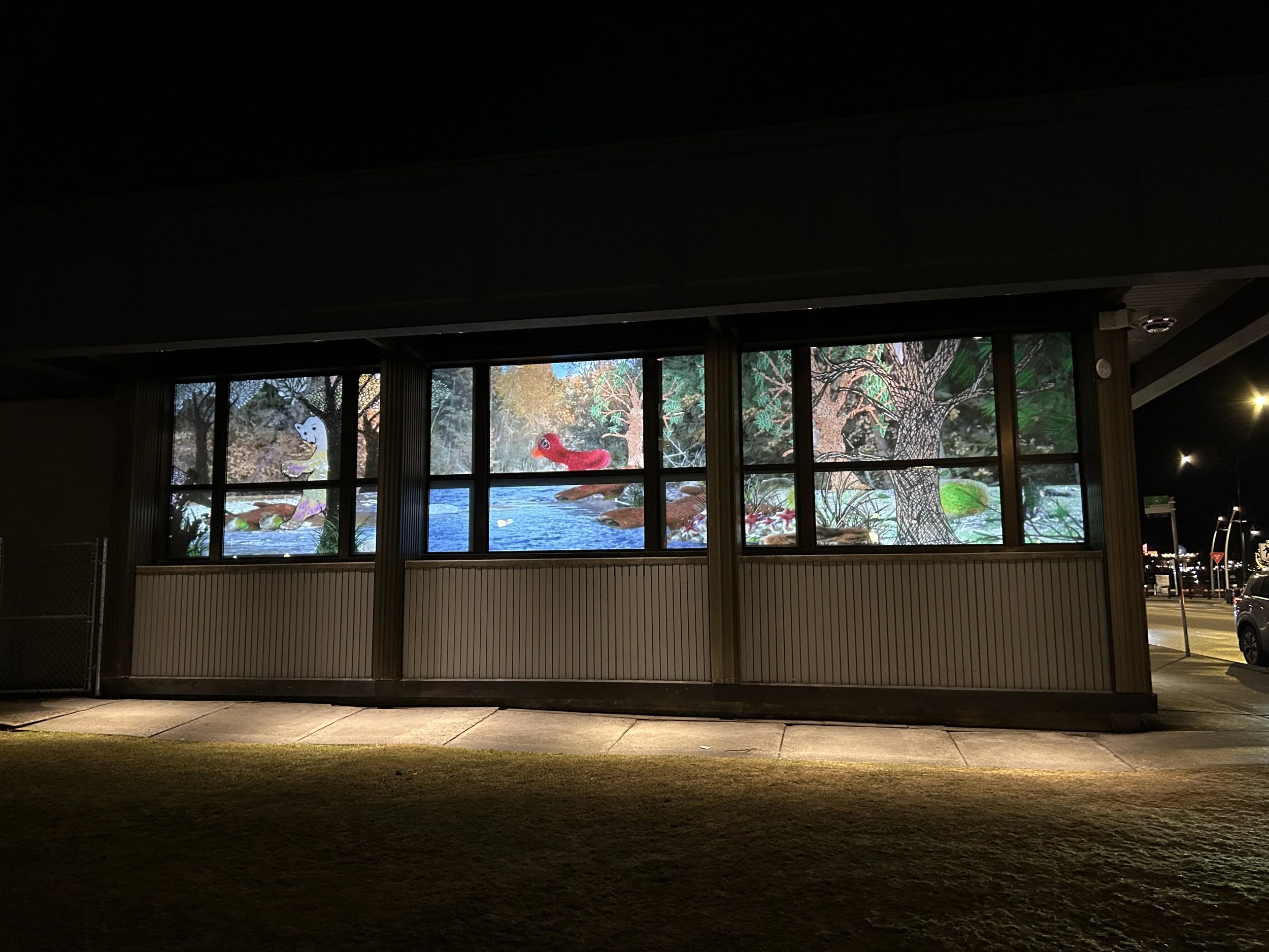 Night scene of a colorful mural on a building's window wall depicting aquatic animals, trees, and a cartoonish snake, illuminated from inside.
