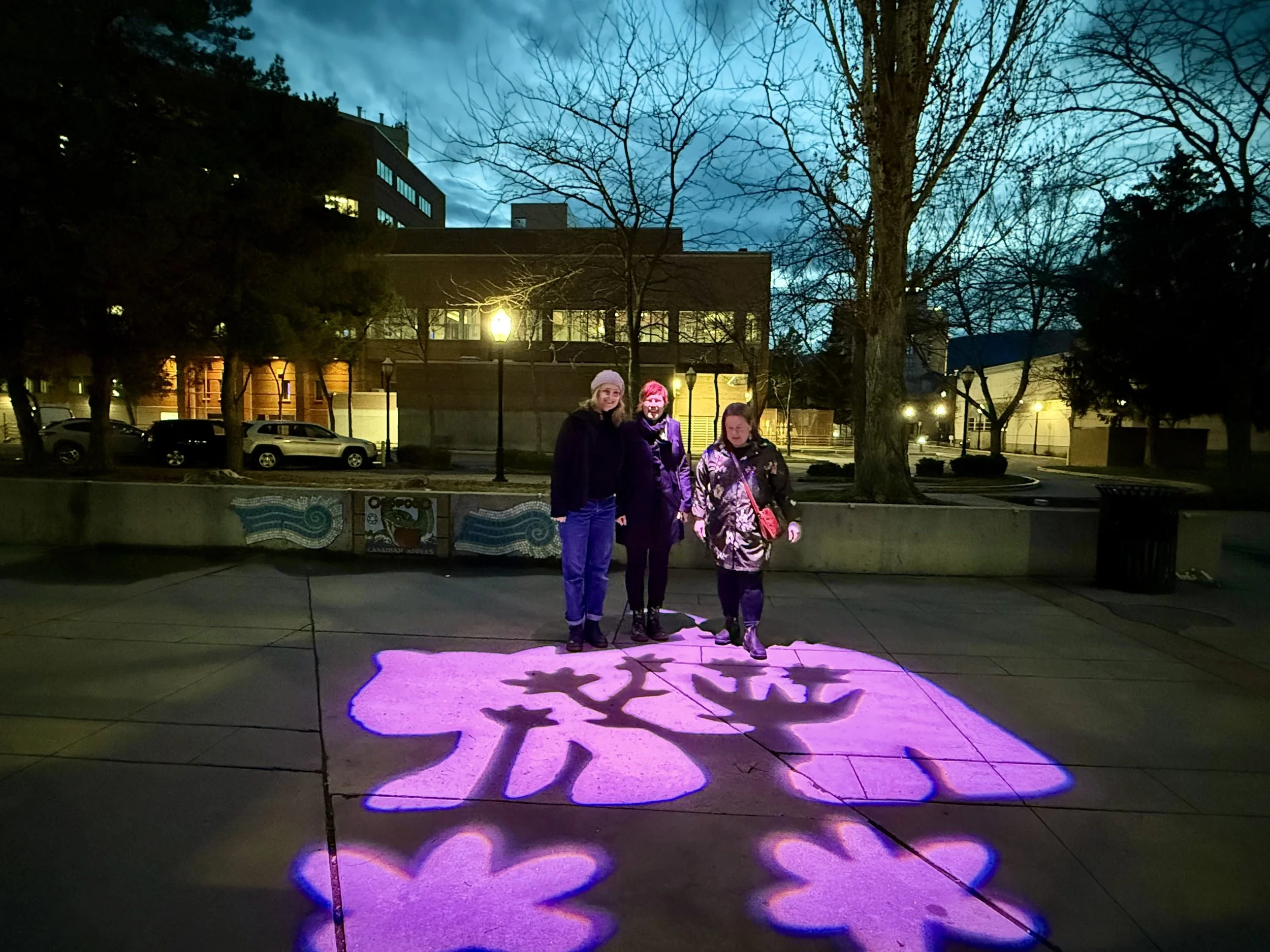 Three women walking on a sidewalk at night with purple blossom shaped projection on the ground, in an urban park area with trees, buildings, parked cars, and lit lamps in the background.