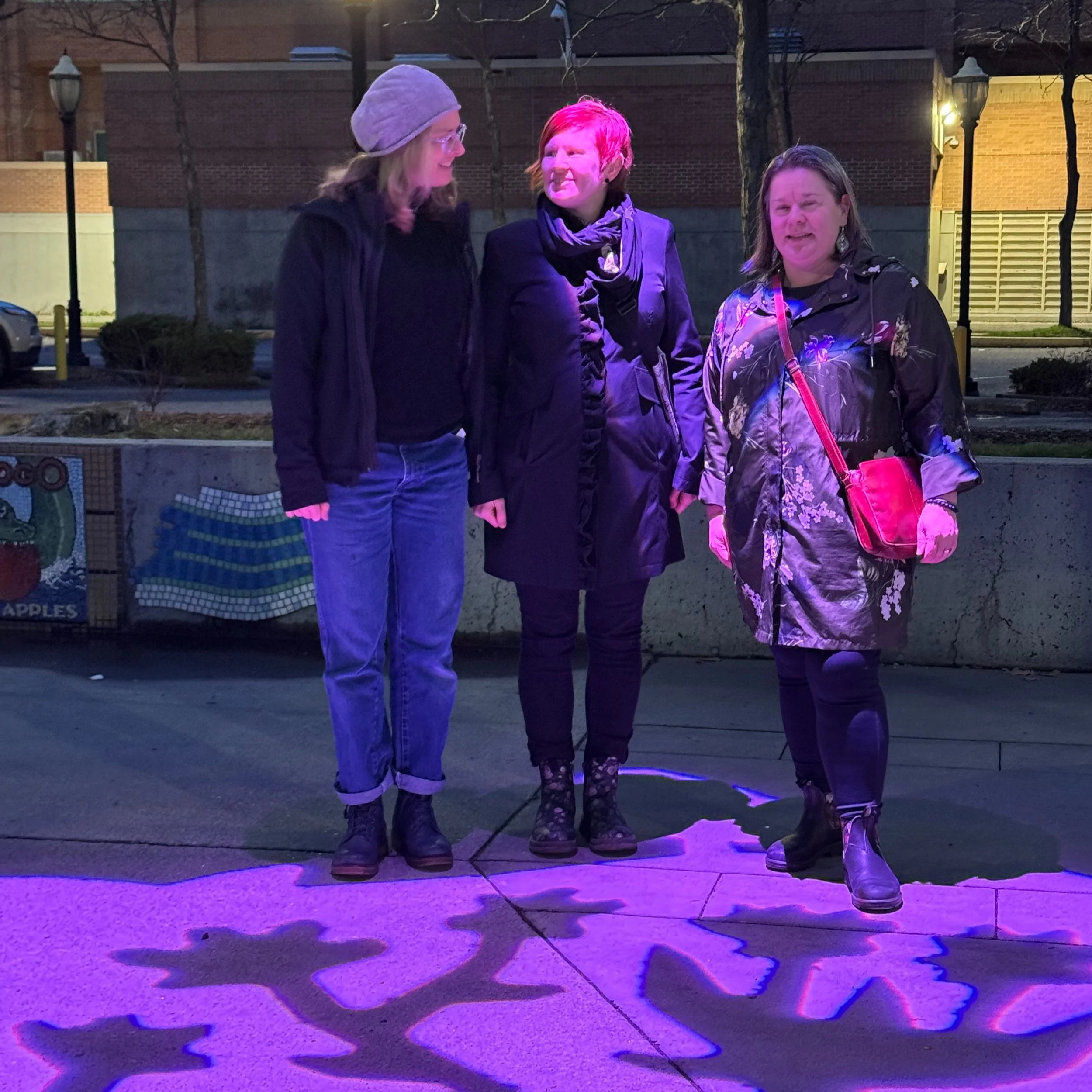 Three women standing outdoors at night with purple lighting, casting shadows on the ground, near a concrete wall with painted artwork.