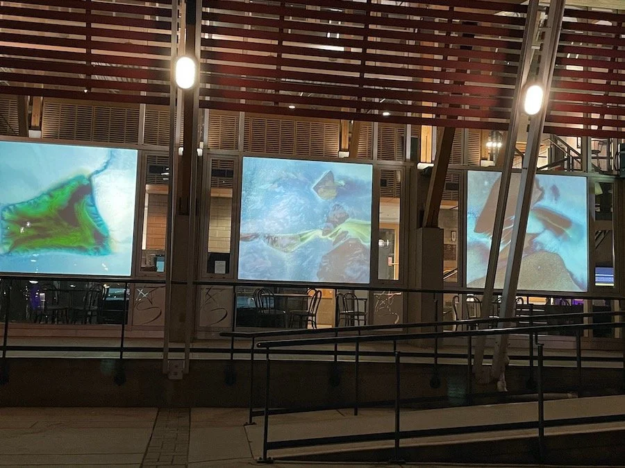 Nighttime view of an outdoor area with three large screens displaying colorful abstract visuals, with tables and chairs visible behind a glass railing.