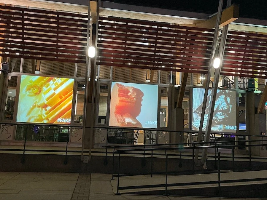 Three digital screens displaying abstract art images with the hashtag #FAKE, set in an outdoor shopping or dining area at night. The screens are mounted behind a railing, and there are overhead lights and structural elements visible.
