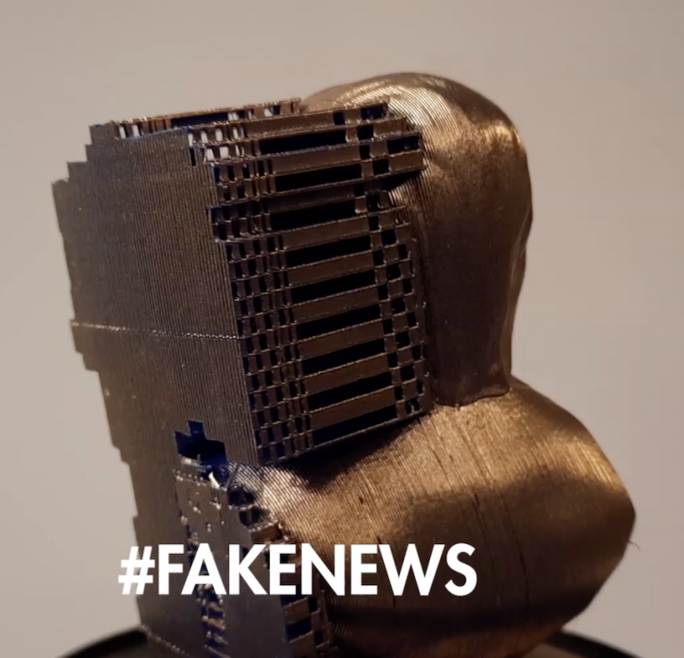 A metallic sculpture of a human head with what appears to be a computer chip or electronic component encased in the forehead, with the hashtag #FAKENews displayed at the bottom.