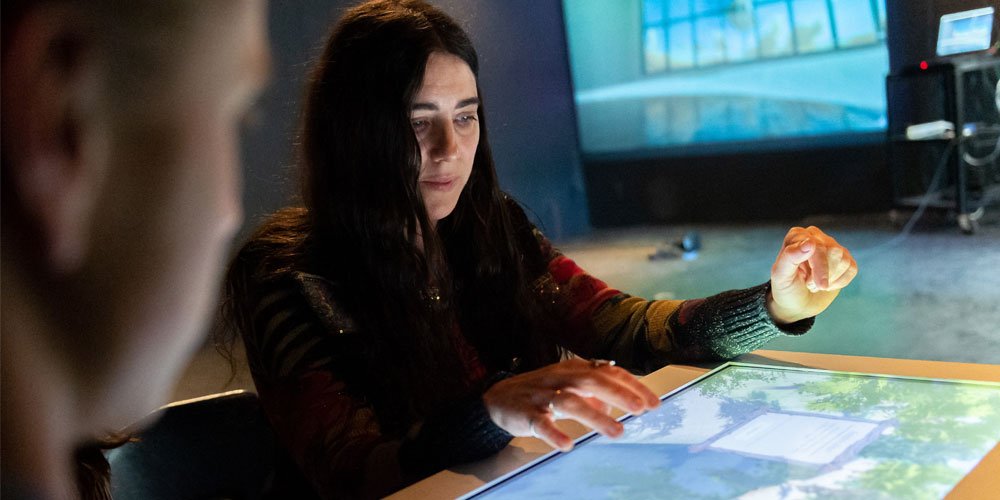 A woman using a large touchscreen table in a dark room, with a blurred person in the foreground.