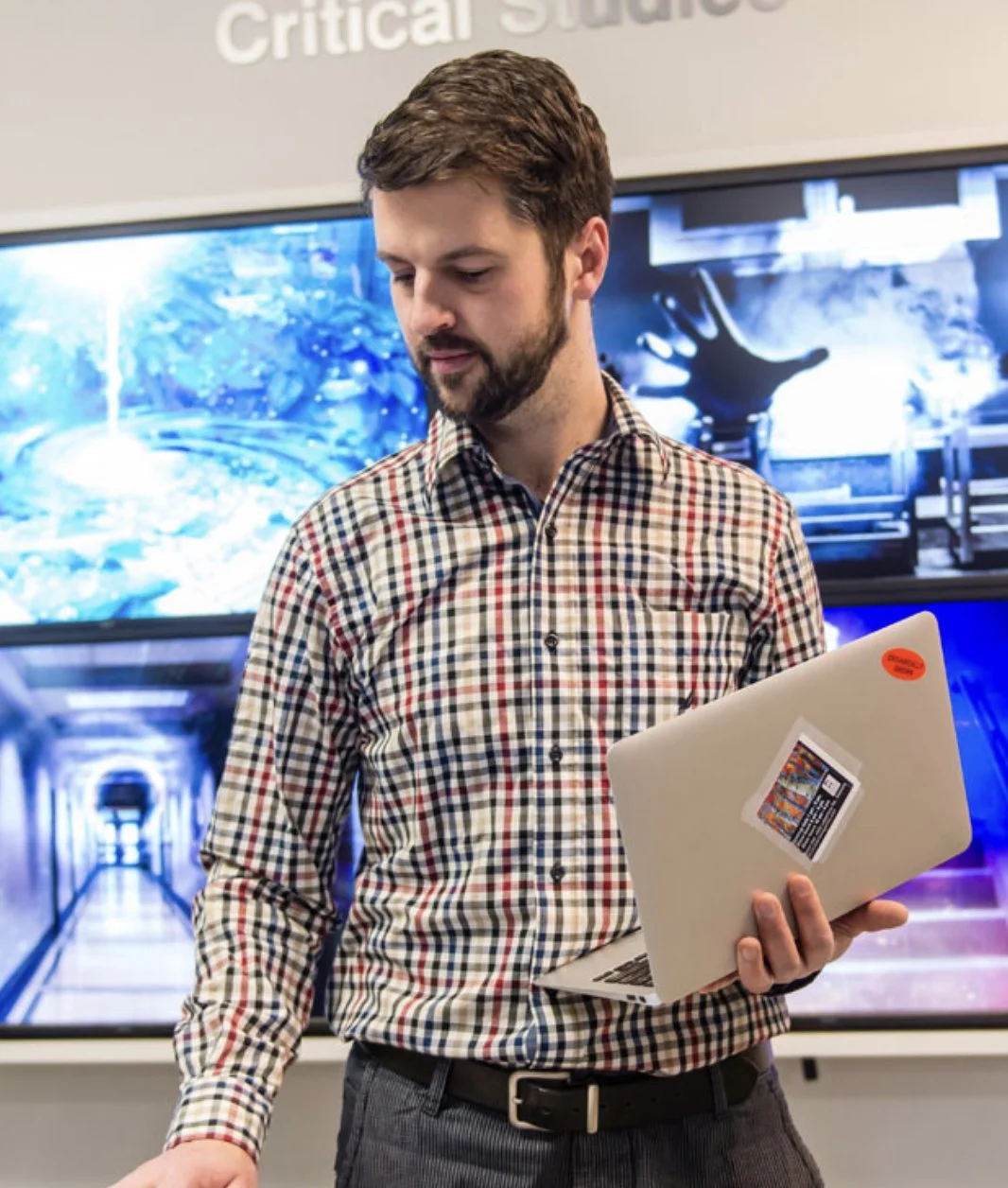 A man with a beard in a checked shirt holding a silver laptop with stickers, standing in front of screens displaying digital art or visual effects.