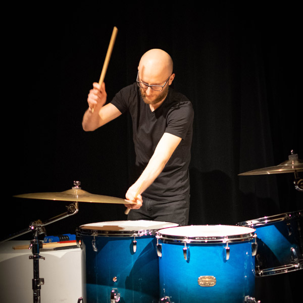 A bald man wearing glasses and a black t-shirt playing drums on stage with a black curtain in the background.
