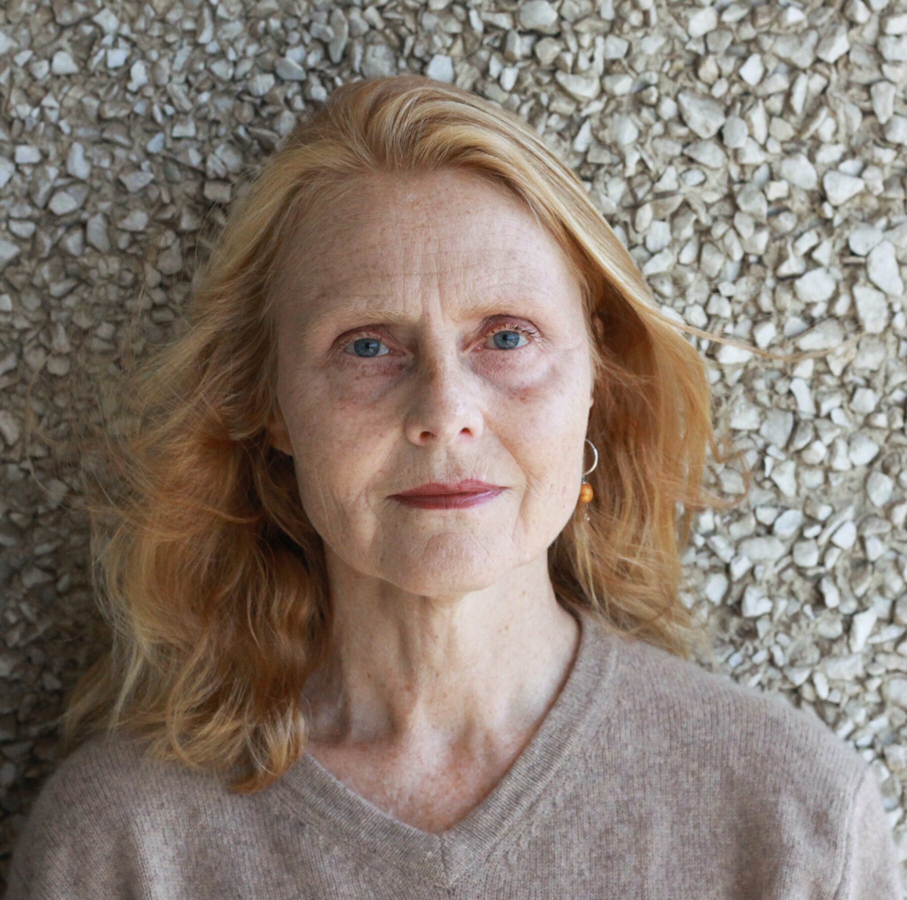 A portrait of a middle-aged woman with red hair and blue eyes, wearing a beige sweater, against a textured stone wall.