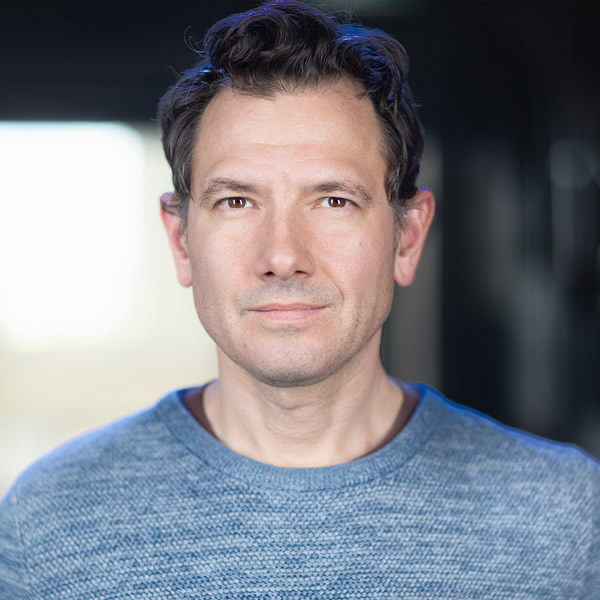 A man with dark wavy hair wearing a blue sweater, looking directly at the camera with a neutral expression, standing indoors.