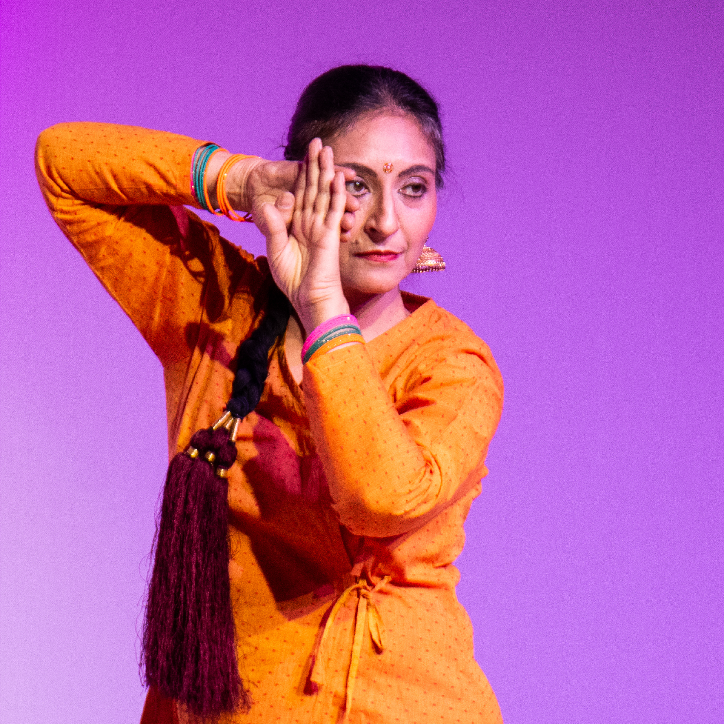A woman dressed in traditional Indian attire with orange dress, jewelry, and colorful bangles, performing a dance pose with her hand near her face against a purple background.