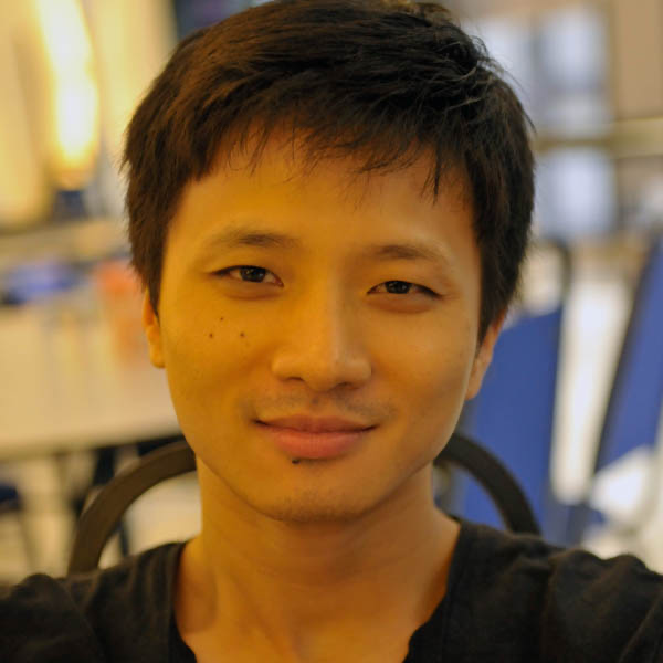 A young man with short black hair smiling, sitting indoors with a blurred background of tables and chairs.