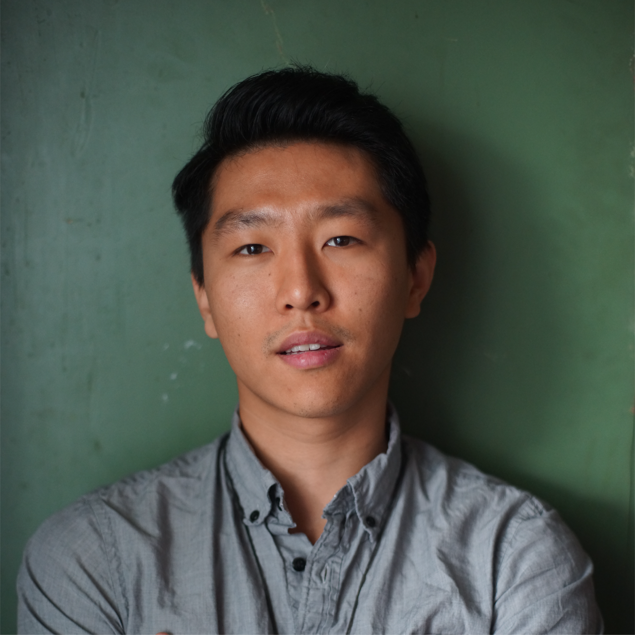 Portrait of a young Asian man with dark hair, wearing a light gray button-up shirt, standing against a green wall.
