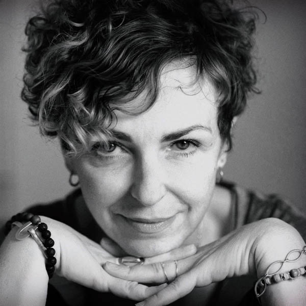 Black and white close-up portrait of a woman with short, curly hair, smiling softly with her hands gently supporting her chin, wearing earrings and bracelets.