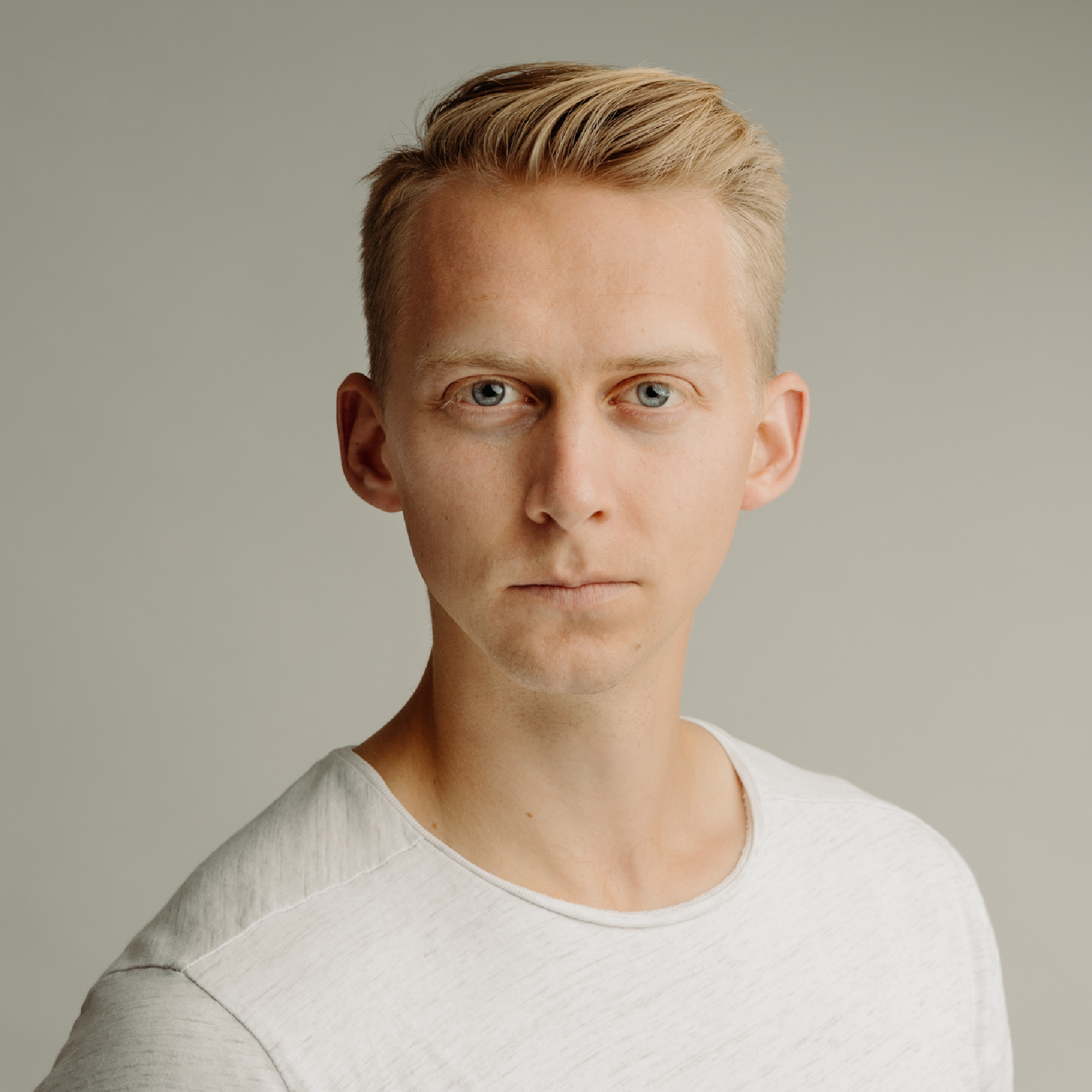 A young man with short blonde hair and blue eyes, wearing a light-colored t-shirt, looking directly at the camera with a neutral expression, against a plain, light gray background.