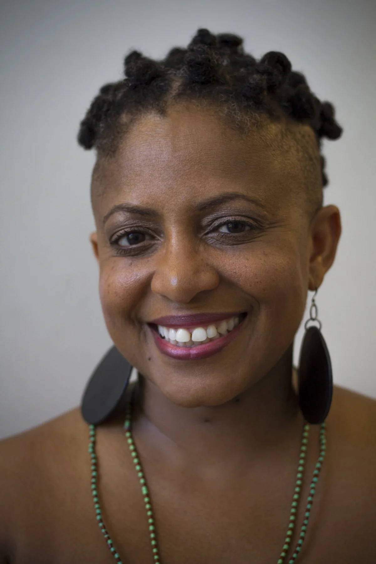 Close-up of a smiling woman with short, textured hair, earrings, and a beaded necklace, against a plain background.