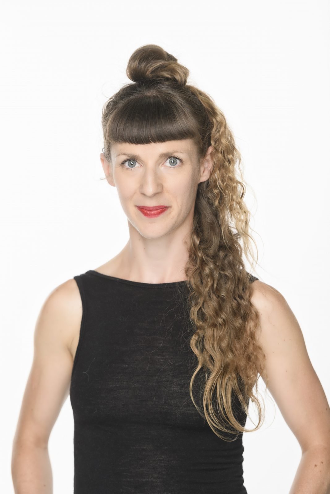 Woman with brown hair styled in an updo with a side ponytail, wearing a black sleeveless top, standing against a white background.